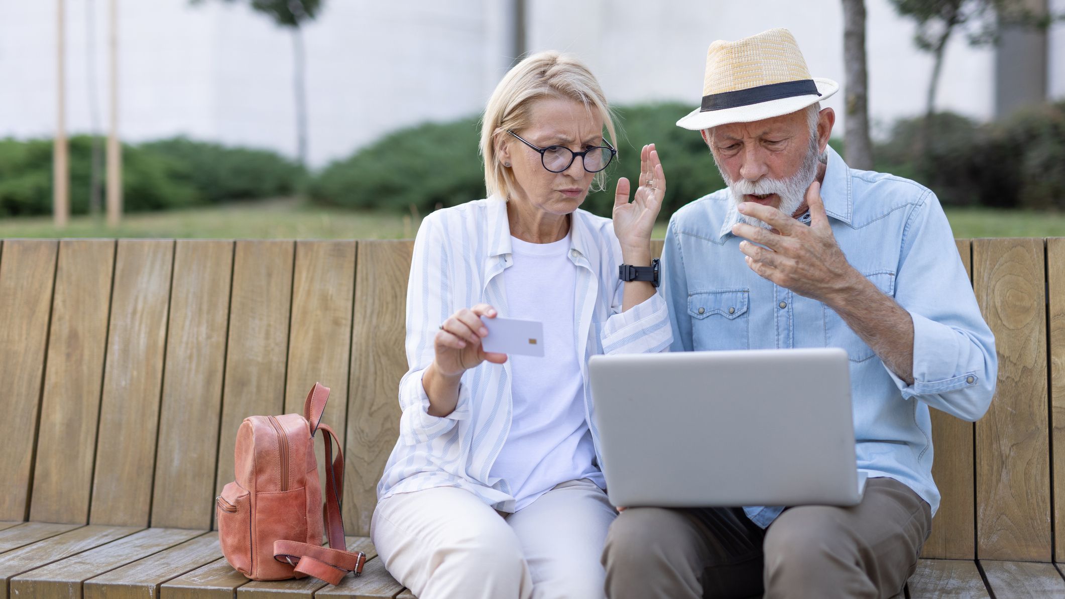 A mature couple sits closely on a wooden bench, showing concern as they look at a laptop, highlighting the reality of dealing with challenges together in an outdoor park setting.
