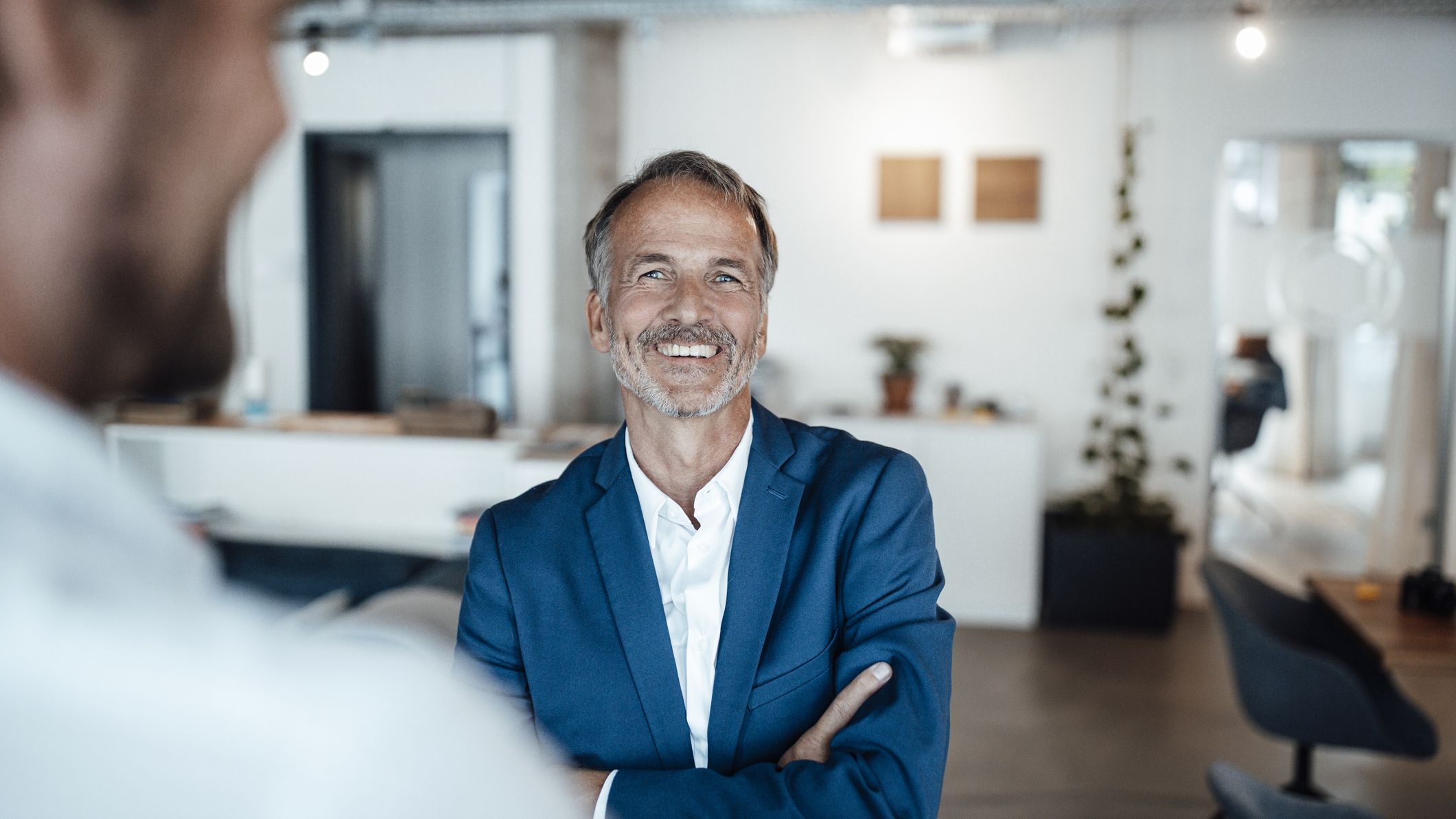 An older man in a blue blazer smiles confidently at the camera. He is talking to a work colleague in the foreground. He looks happy to be at work.