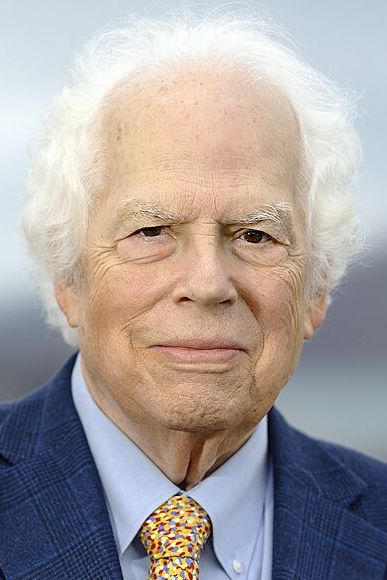 A man with wavy white hair wearing a blue blazer, blue button up and yellow-patterned tie looks at the camera