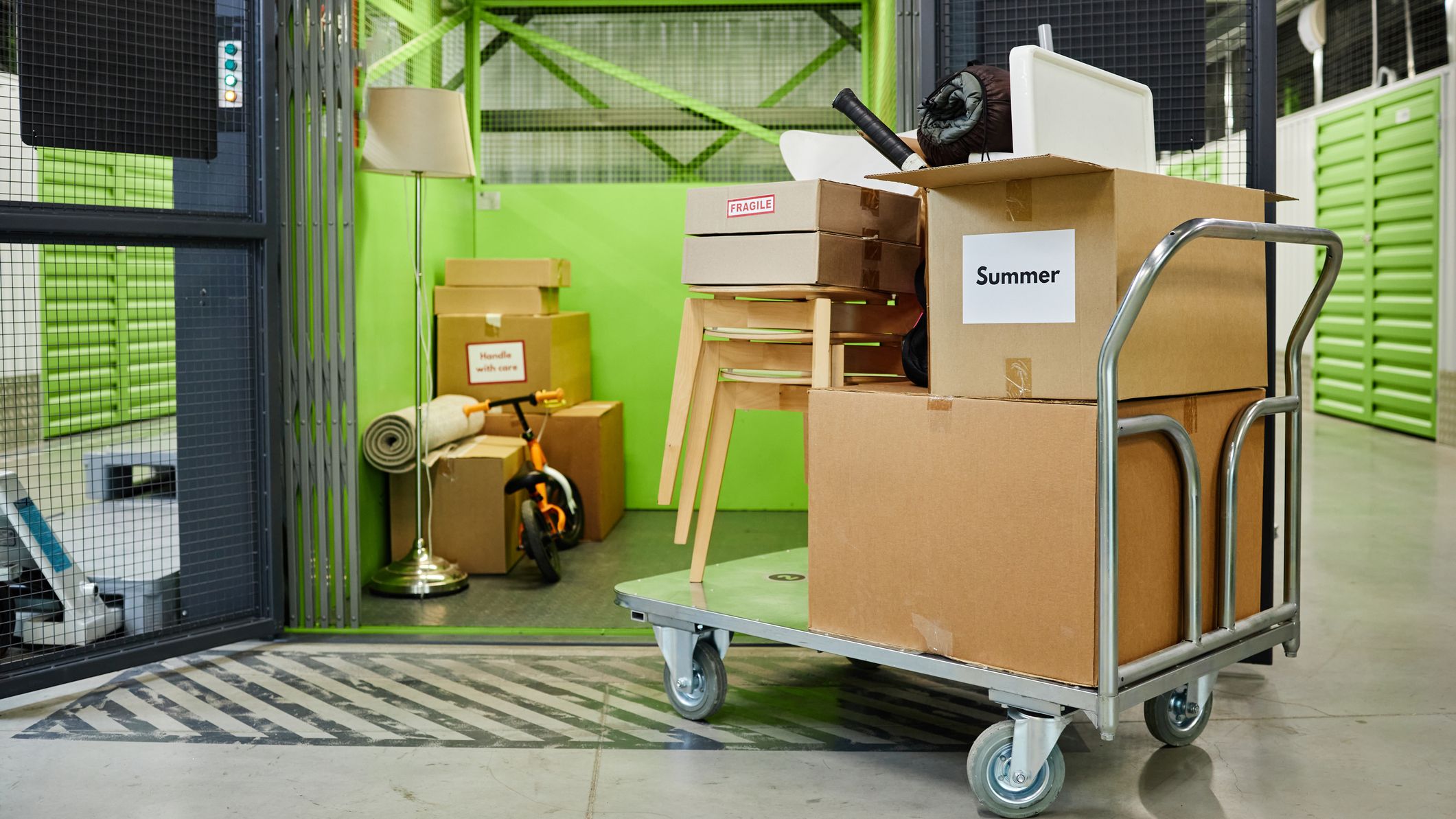 Stacked cardboard boxes and household items including lamp, chair, and scooter being moved on metal cart in self storage facility with green storage unit in background
