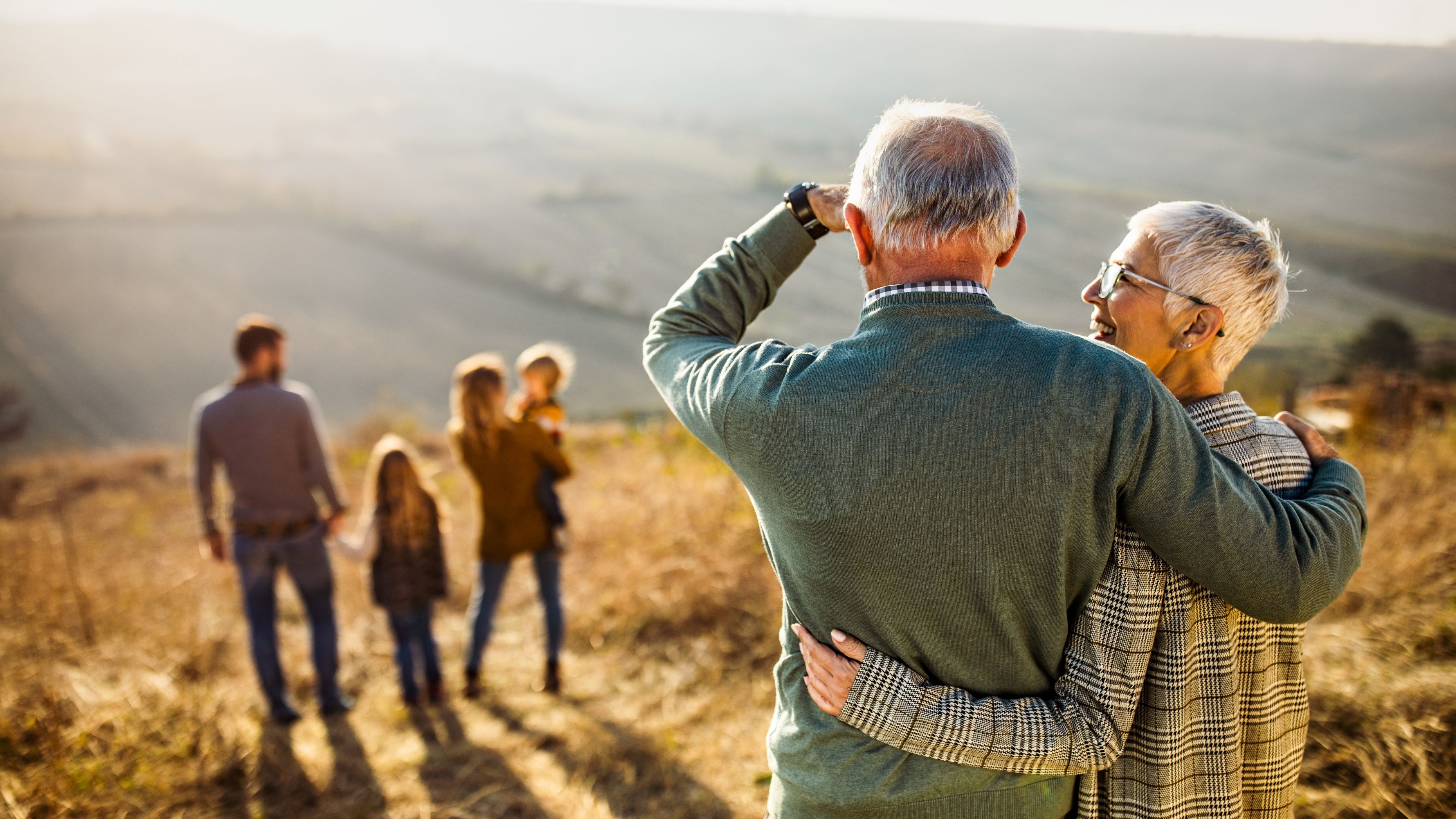 Grandparents stand together while smiling at their family in the distance.