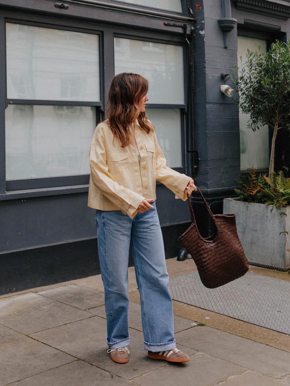 Woman wears cream jacket, blue jeans, brown trainers, knitted leather bag