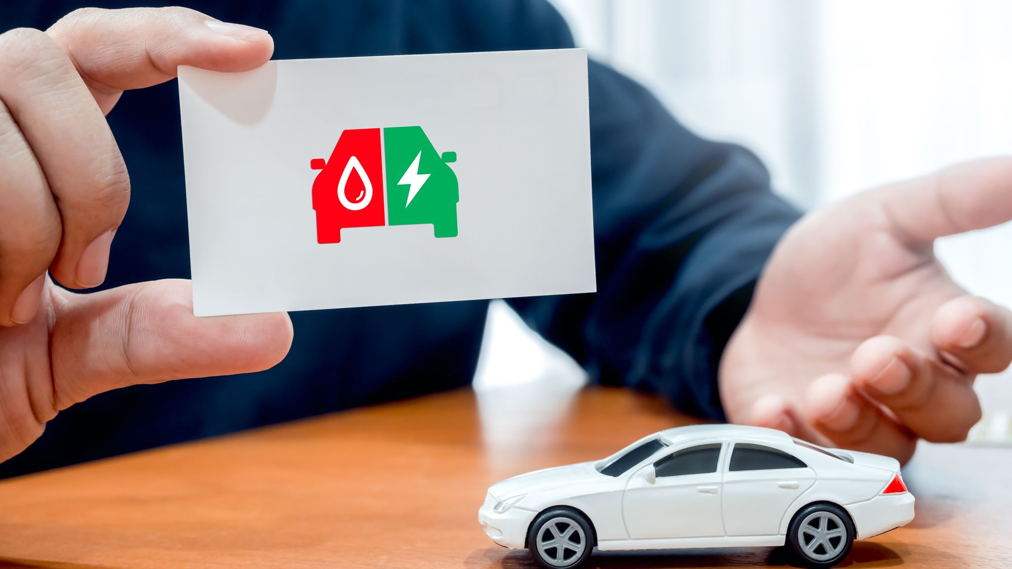 Electric energy and fuel engine, plug-in hybrid electric vehicles, on white business card in broker or agent hand near car toy on wood table.