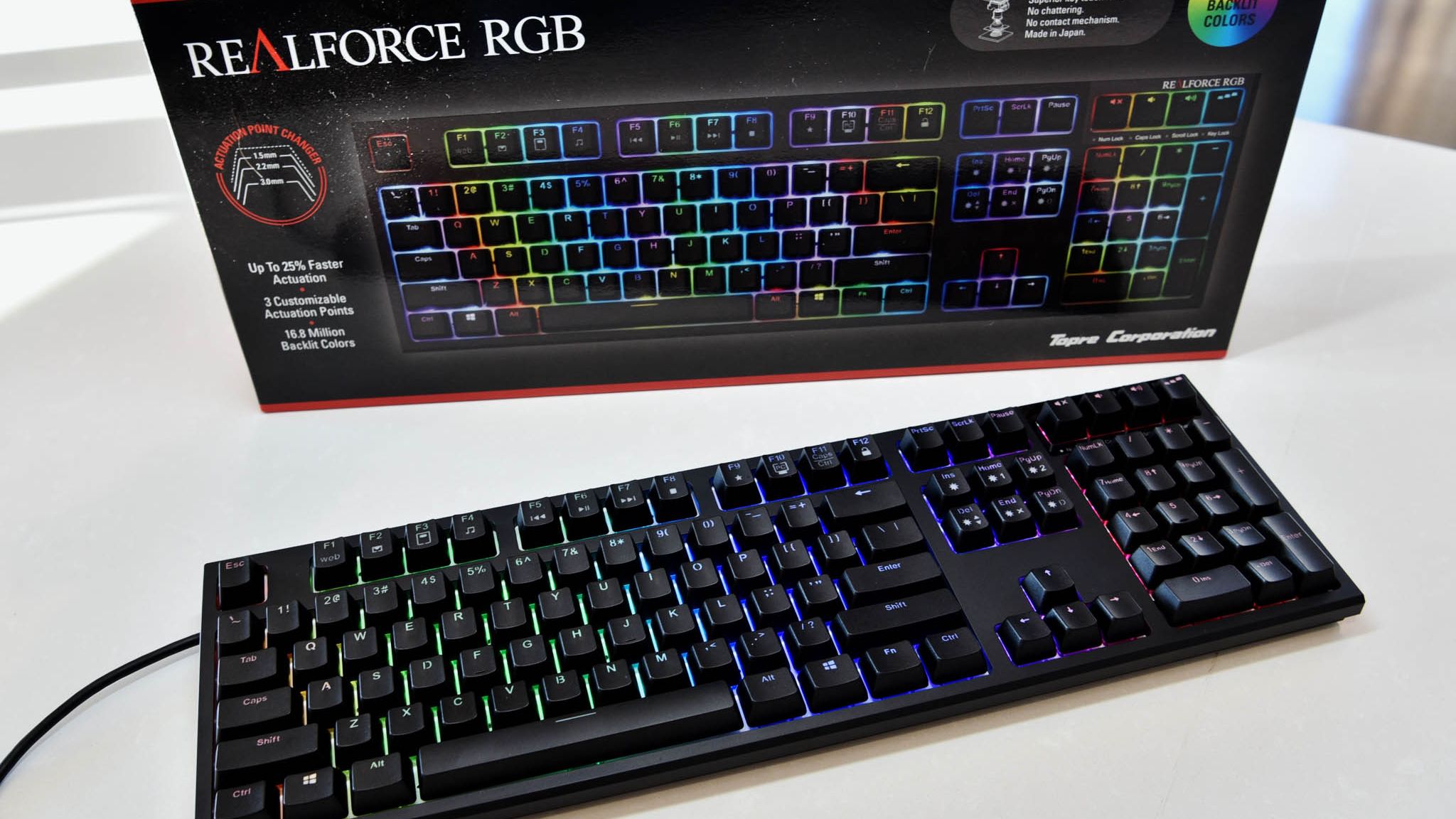A RealForce RGB keyboard sitting on a white table, with keys backlit in various colors. The product box sits upright above the keyboard.