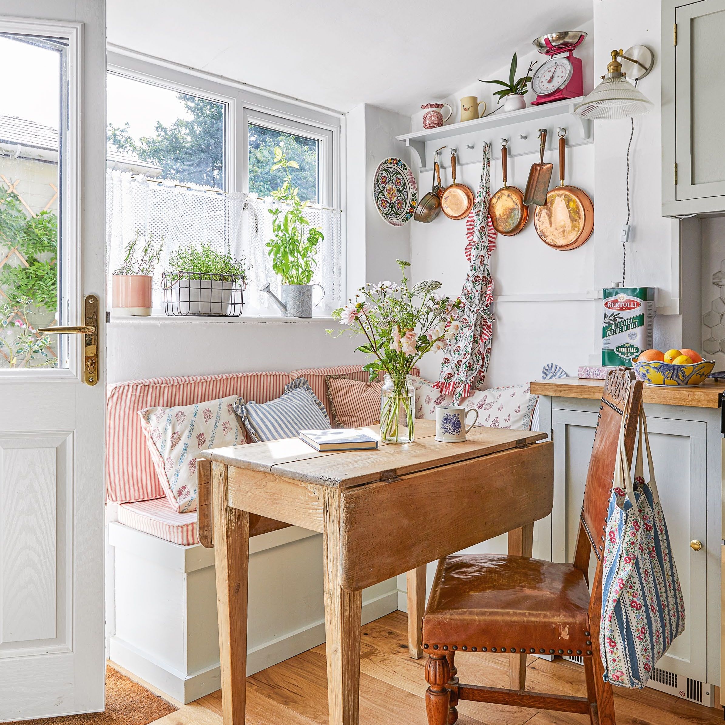 Small kitchen diner area in the corner of a cottage kitchen with drop-leaf wooden table, banquette seat and copper pans on wall