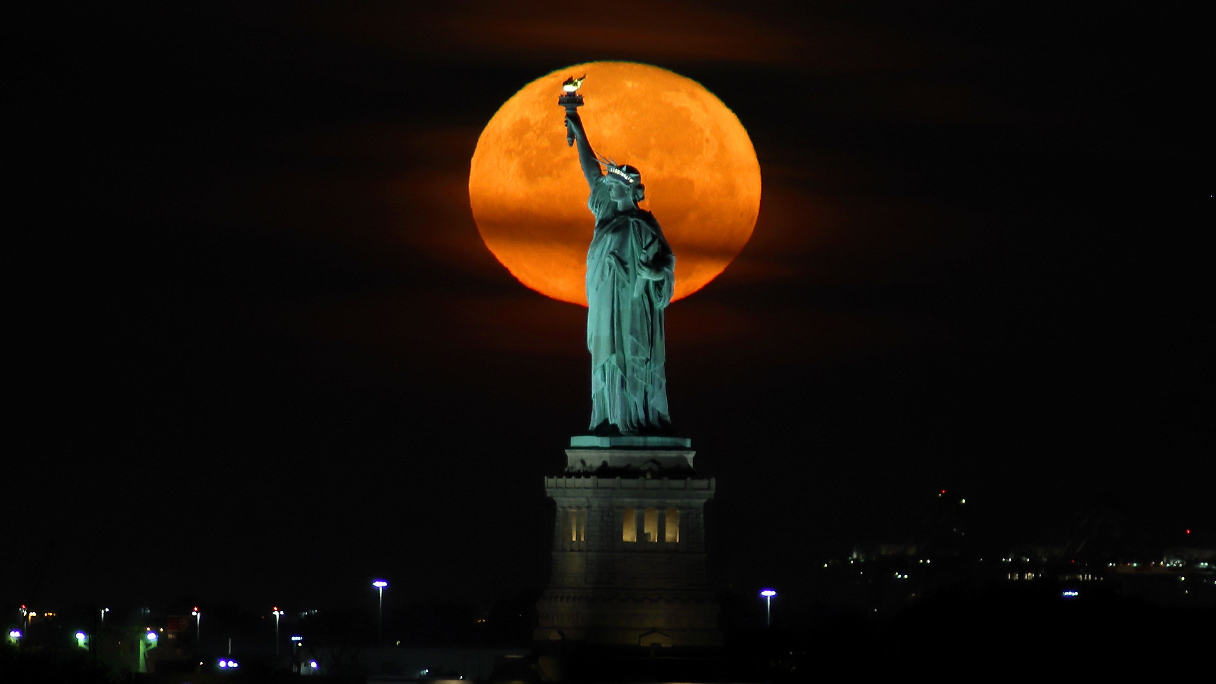 An orange full moon rises behind the Statue of Liberty in New York.