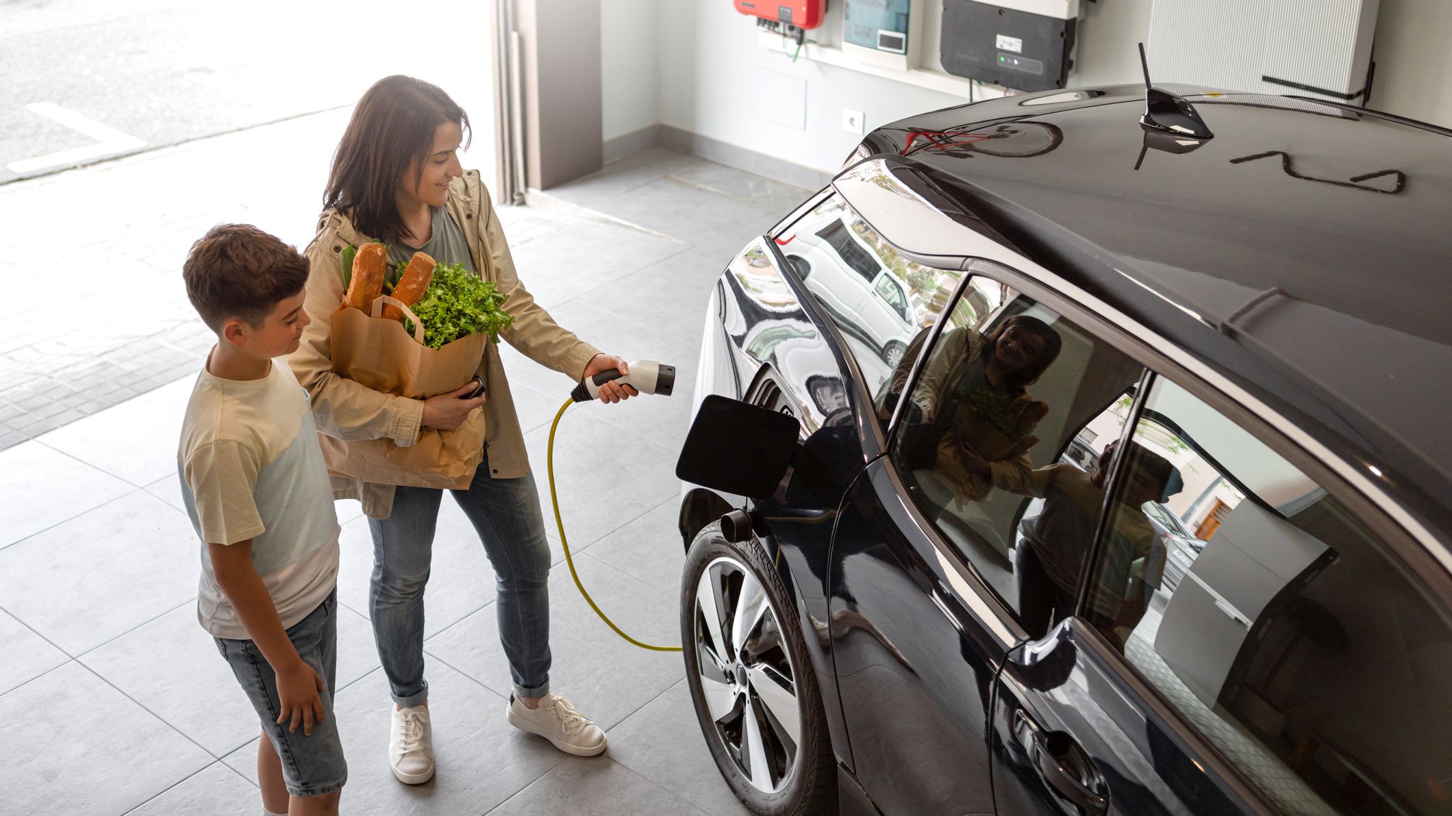 Happy son and mother plug EV charger from home charging station to electric vehicle. Future eco-friendly car powered by renewable source of clean energy on daytime. Horizontal, high angle.