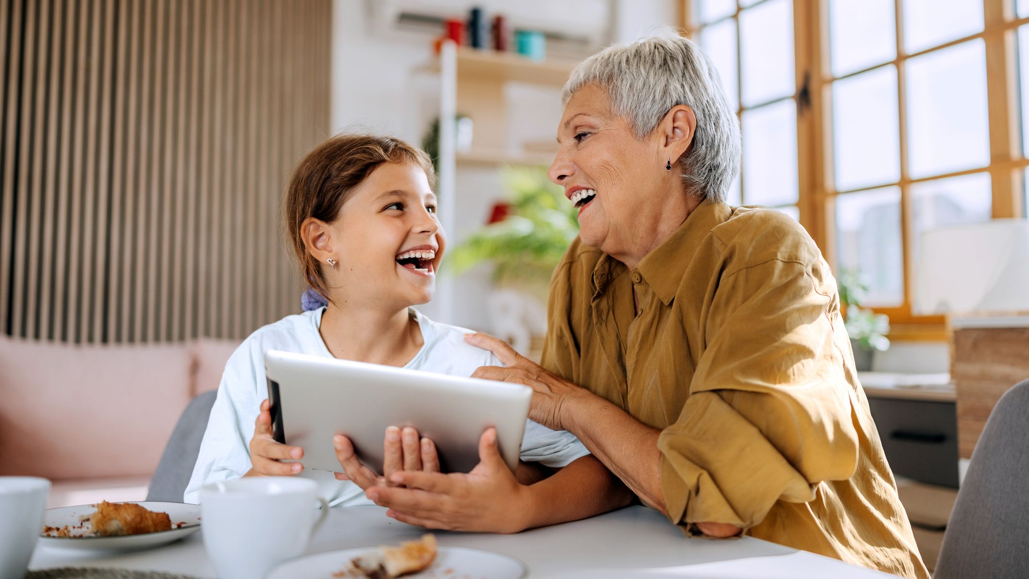 Cheerful grandmother and her young granddaughter sitting at the breakfast table, interacting with a digital tablet. Represents family bonding, technology use, and joyful multigenerational moments.