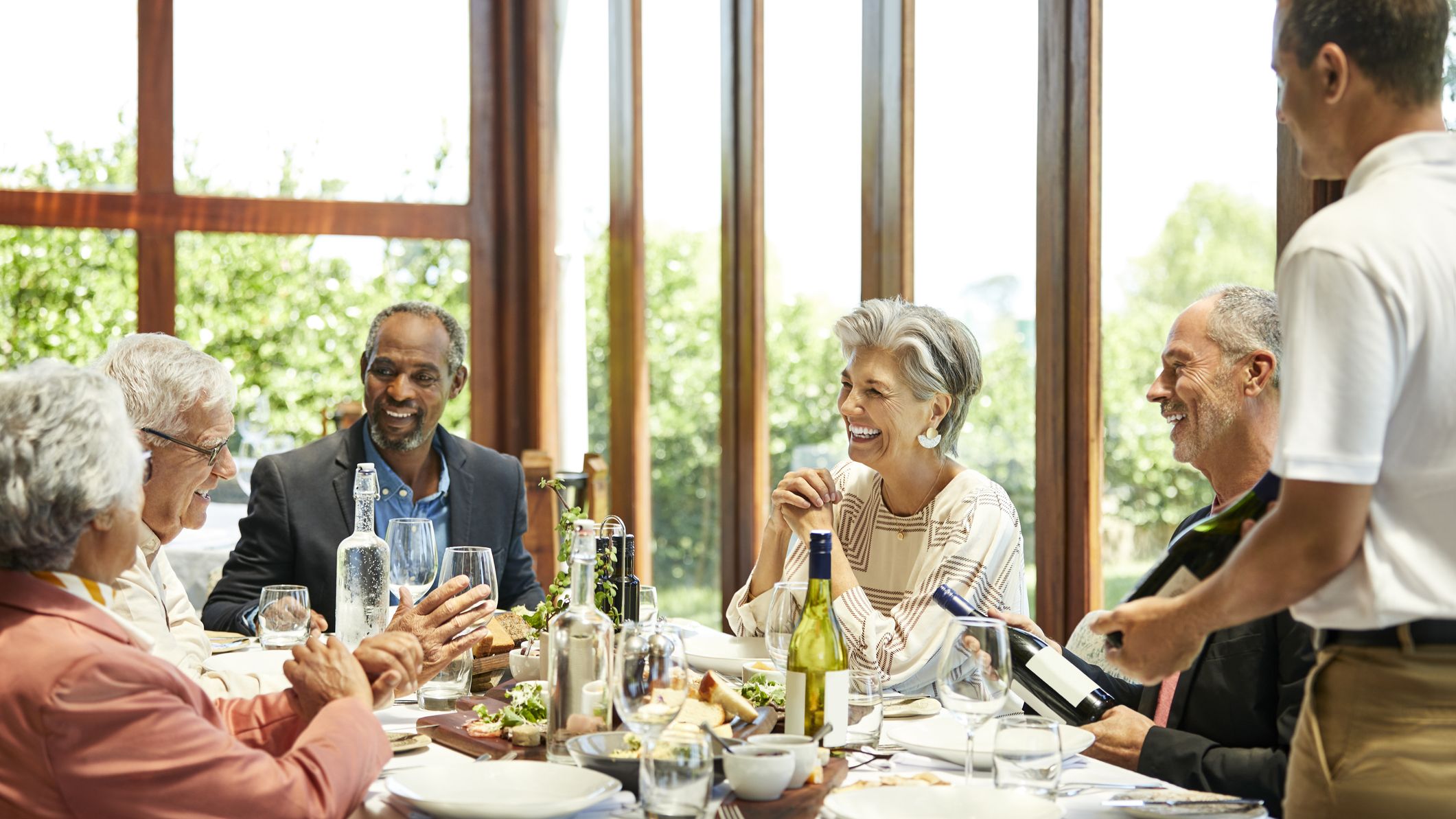 Male sommelier with wine bottle standing by smiling senior friends. Men and women are sitting in restaurant. They are spending leisure time.