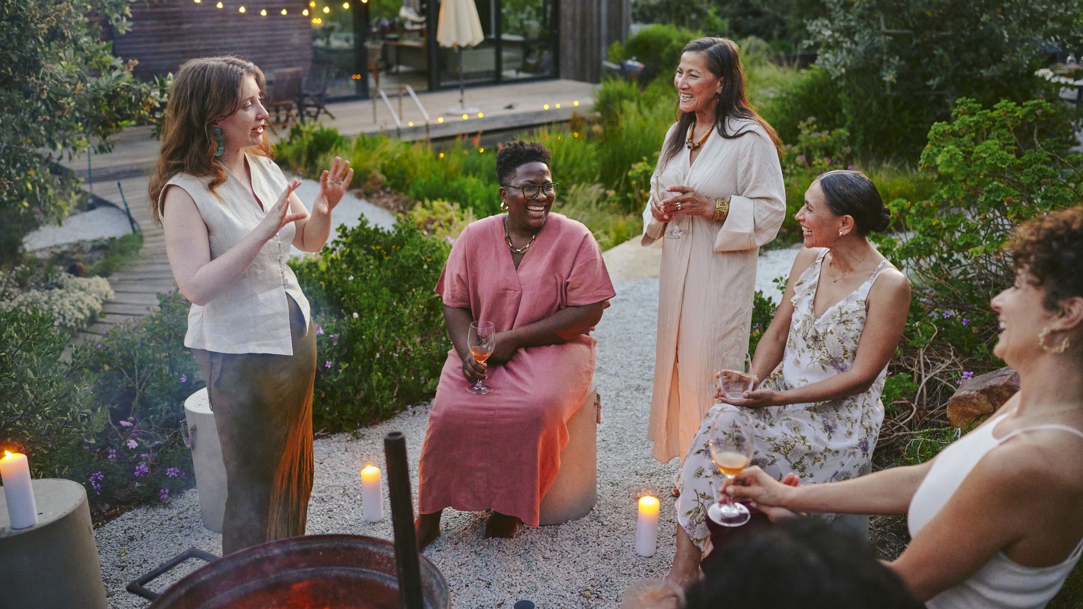 Diverse women laughing over drinks around a fire pit during a garden party