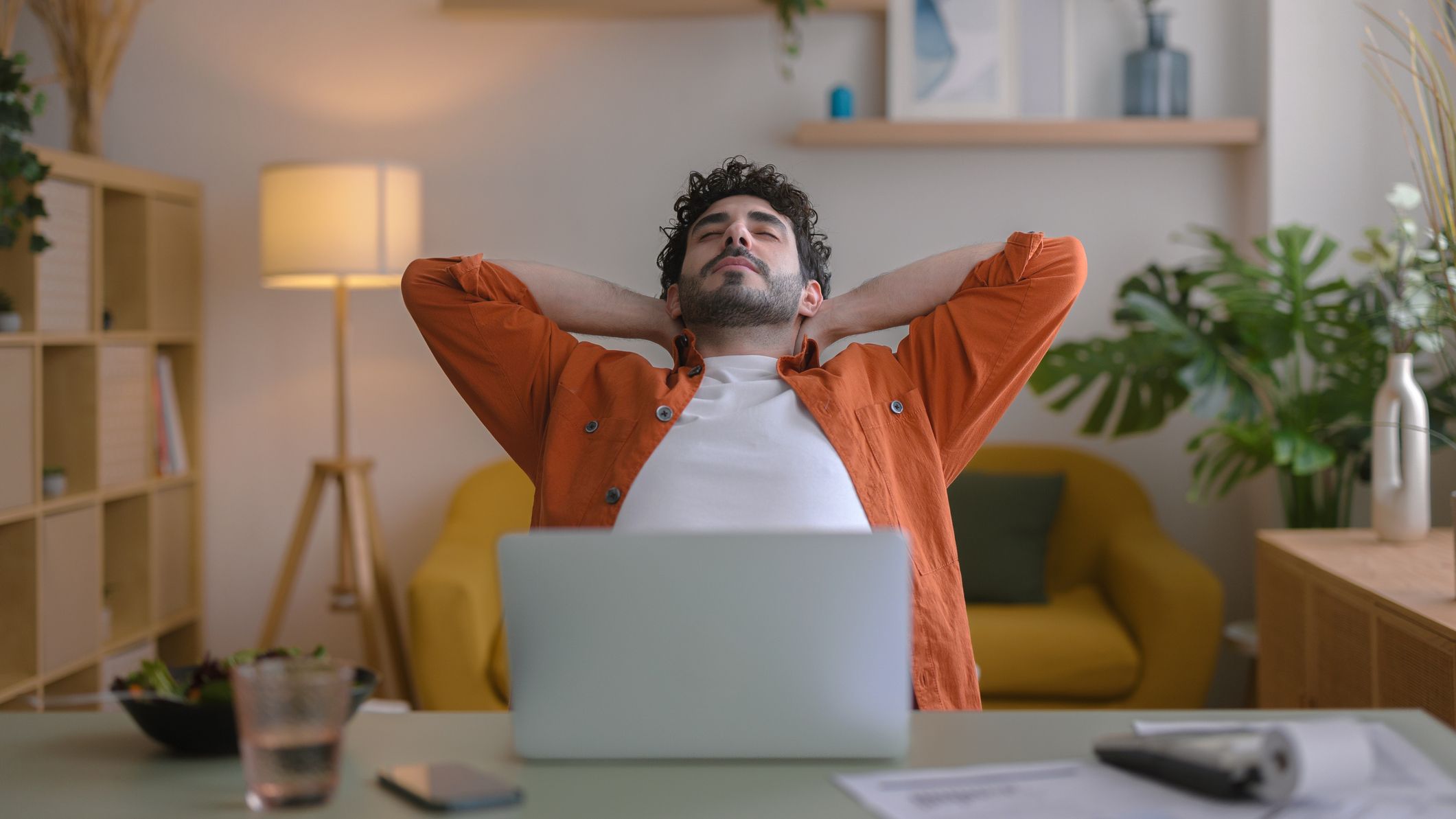 Man sitting in front of a laptop in a domestic setting, leans back with his hands behind his head and his eyes closed