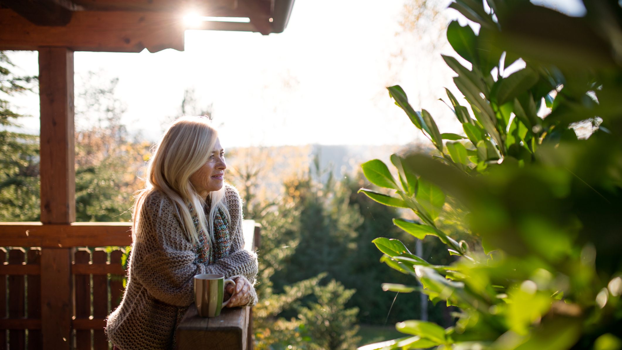 An older, attractive woman smiles and leans on a porch railing. She has a warm cup of tea or coffee and the sun is rising or setting. She exudes calm.