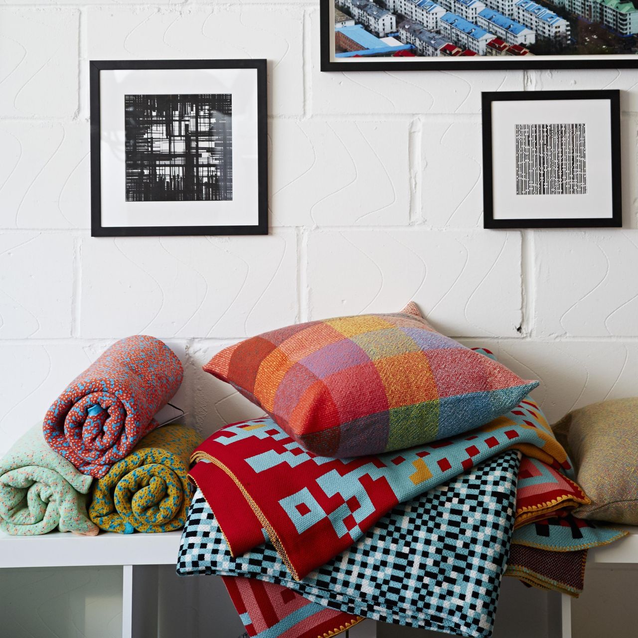 A pile of stacked and rolled colorful blankets against a white wall with artwork above