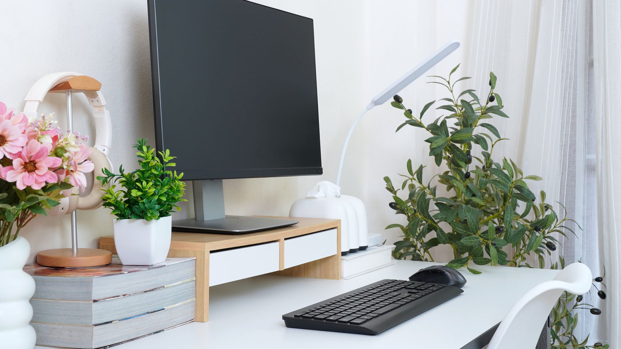 Minimalist home office setup with monitor, keyboard, headphones, books, and potted plants.