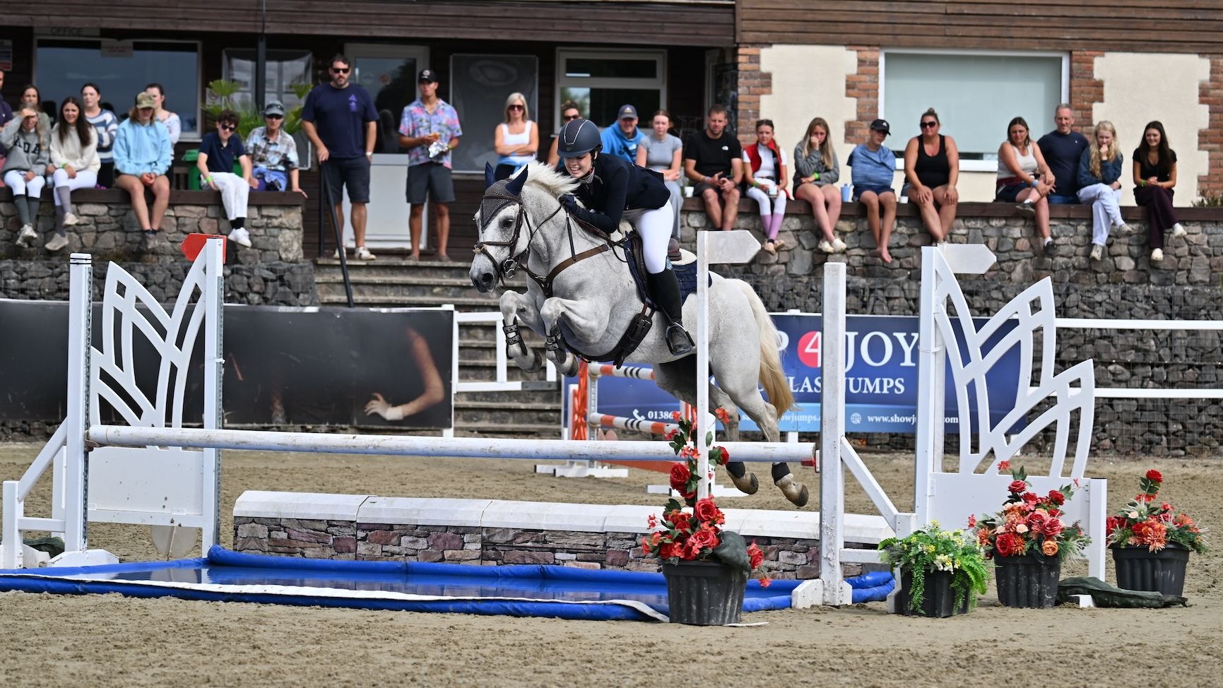 A young rider and a grey pony jumping an open water jump