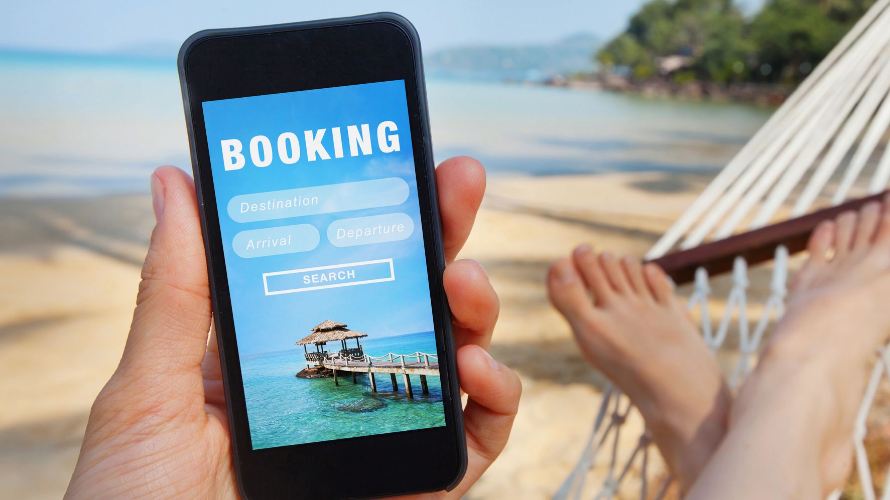 A person looks at a phone with a travel "booking" screen while lying on a hammock on a beach.