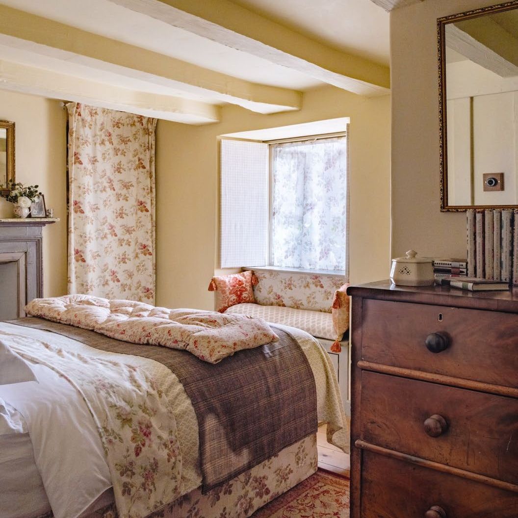 Cottage bedroom filled with floral fabrics, with a window seat and antique chest of drawers