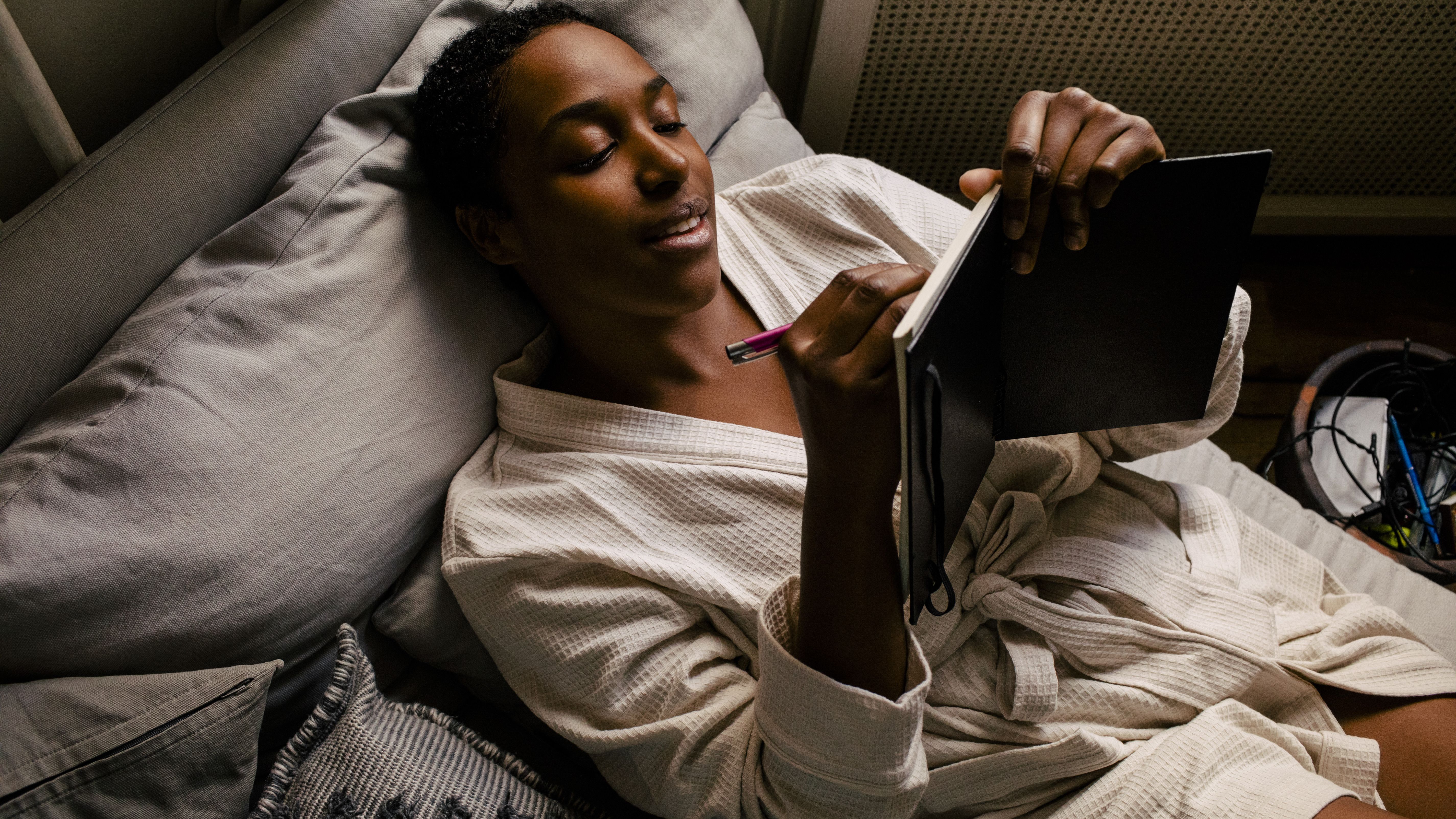 A woman lies in bed wearing a dressing gown as she writes in a journal.