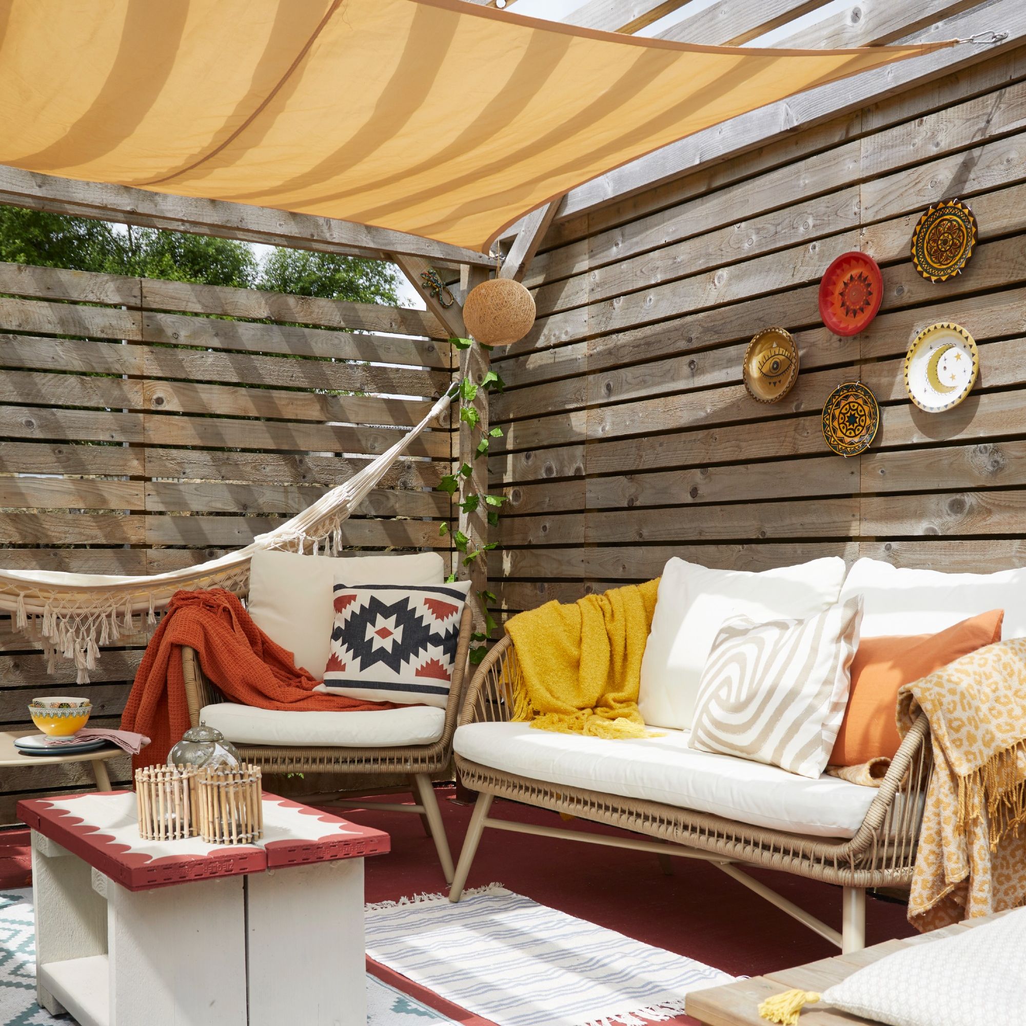 An outdoor living room covered with a pergola and adorned with cushions, throws and rugs on the floor