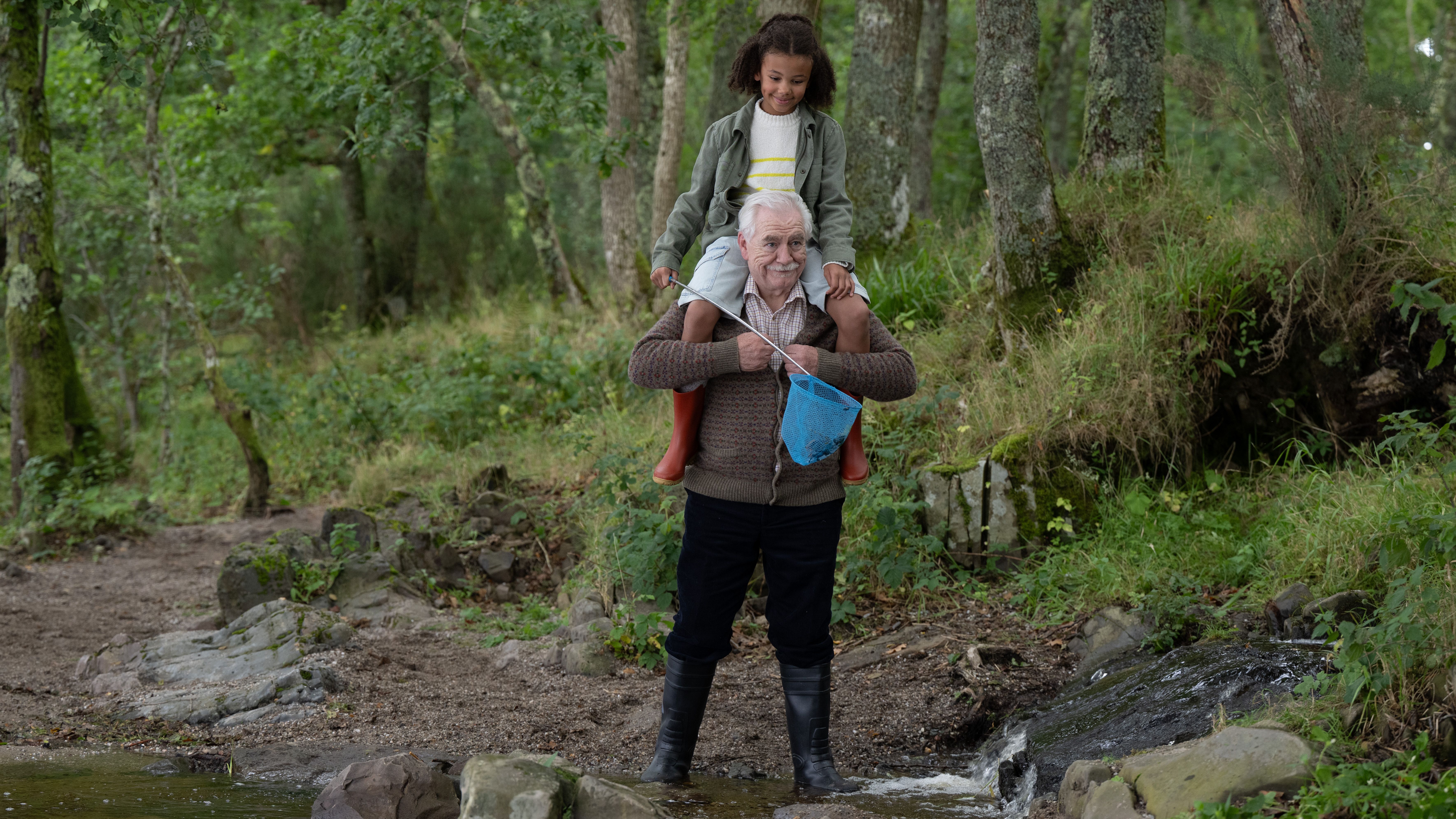 an image of brian cox in character in Glenrothan walking beside a river with his grand-niece on his shoulders