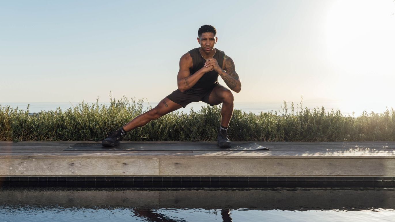Man demonstrates side lunge exercise outside, he is reflected in a body of water in front of you. There is grass and blue sky behind him.