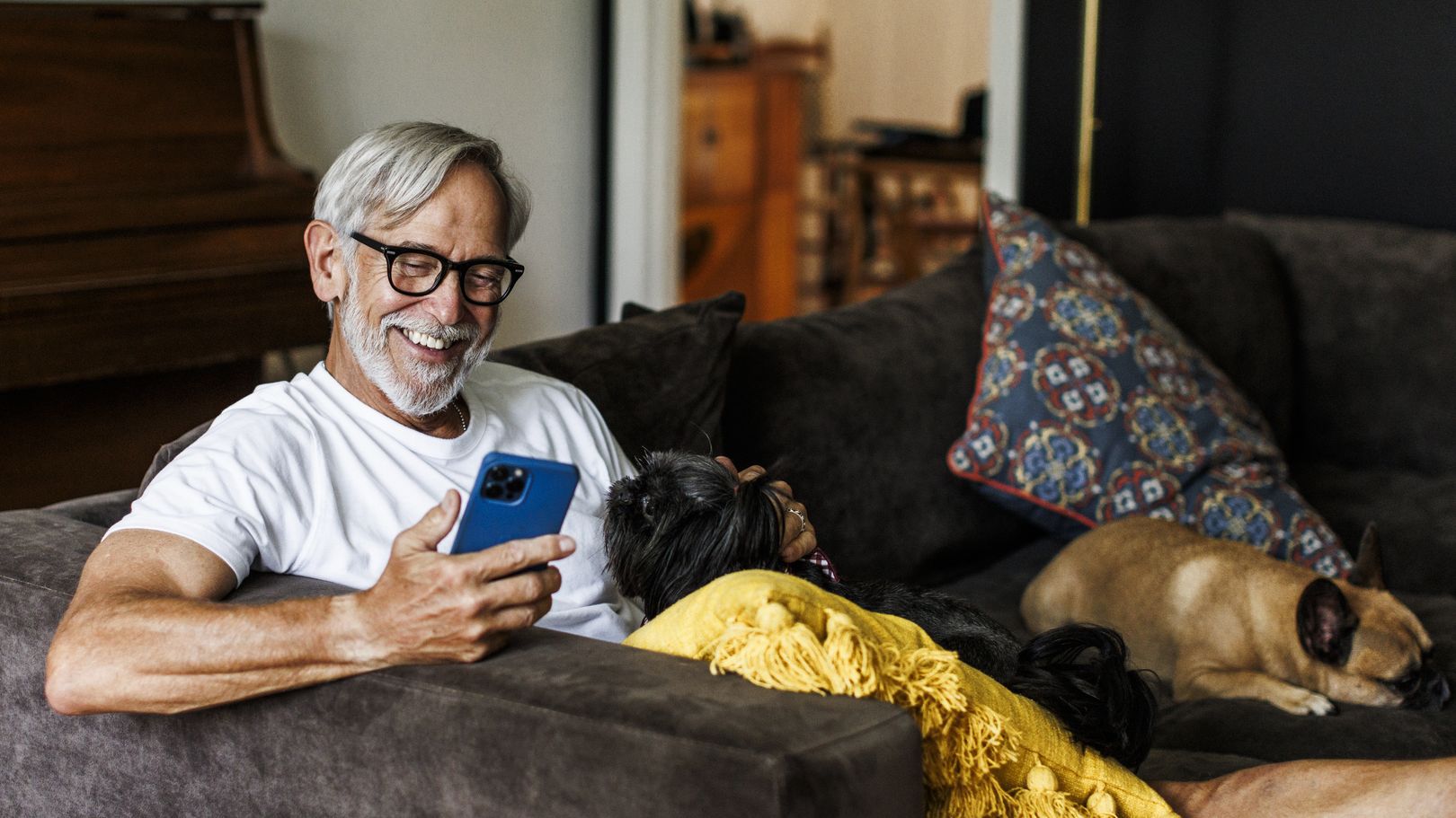 A man relaxing on a sofa at home with his two dogs, reading something on his smartphone.