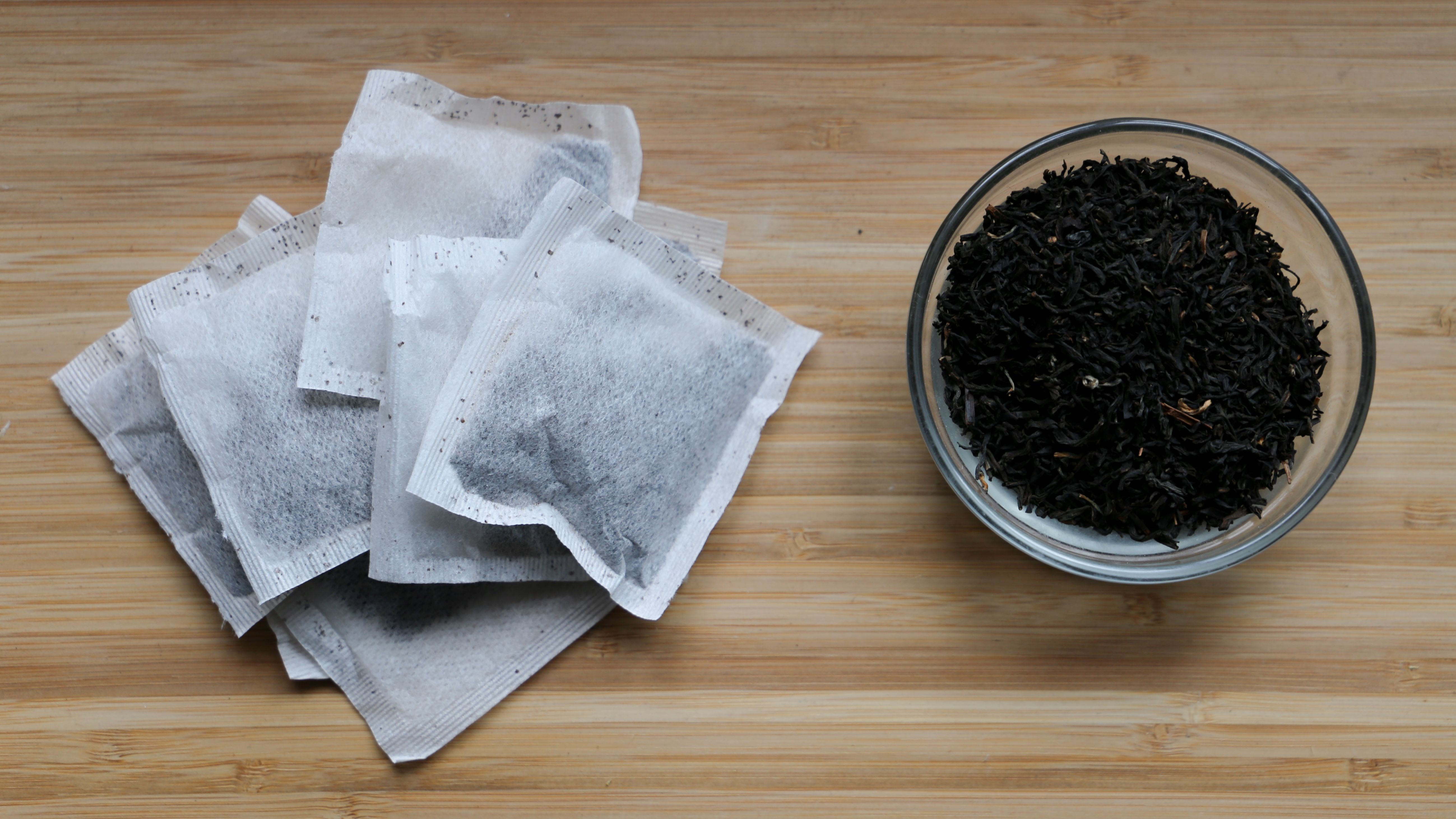 Pile of tea bags on a wooden surface next to a bowl of loose leaf tea.