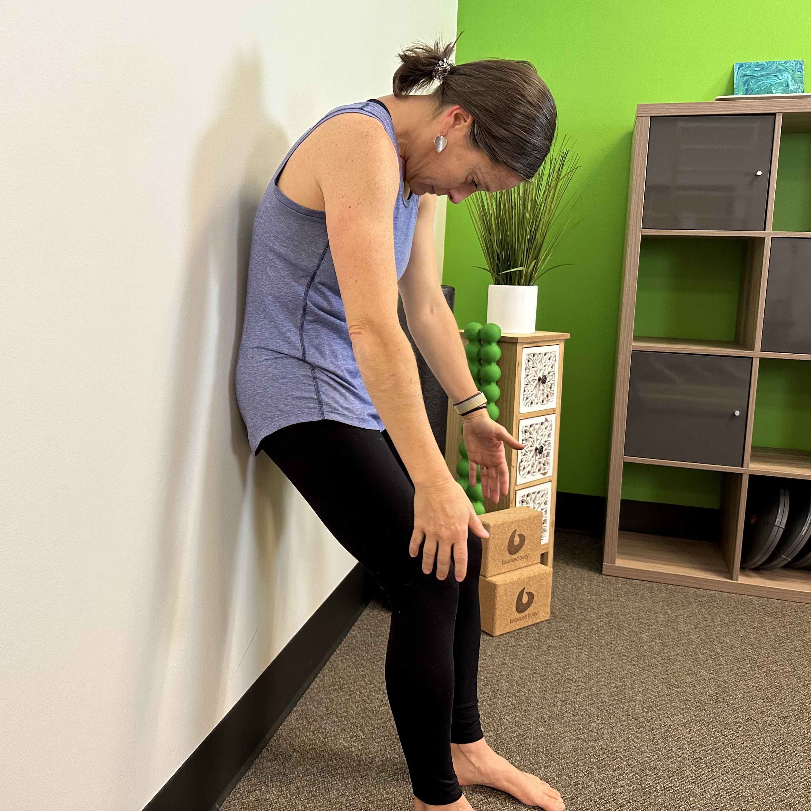 woman in a blue vest and black leggings with her back against a wall half way through a roll down.