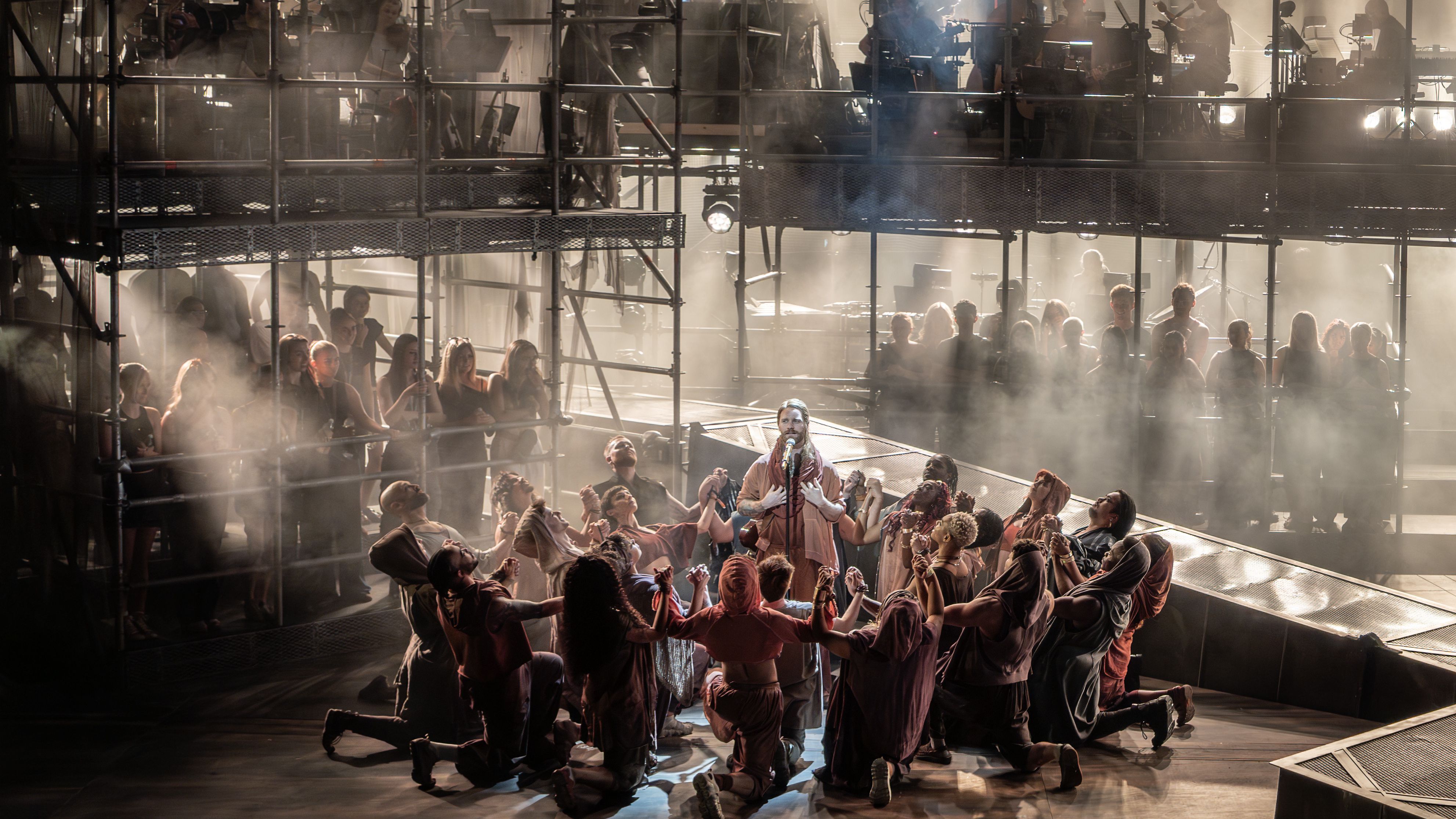 Jesus Christ Superstar musical showing the ensemble all onstage surrounding Sam Ryder in a circle. You can see the orchestra on the scaffolding stage above them, and some audience members to sides