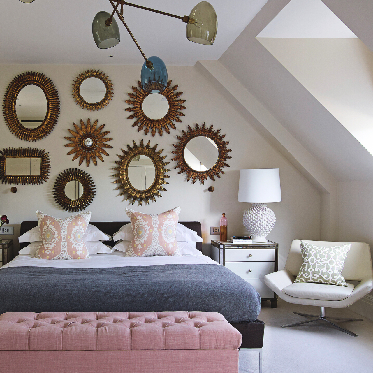 a bedroom with a gallery wall of different sizes of mirrors, a bed with pink, white, and gray bedding, a pink ottoman at the foot of the bed, and an accent chair to the right side