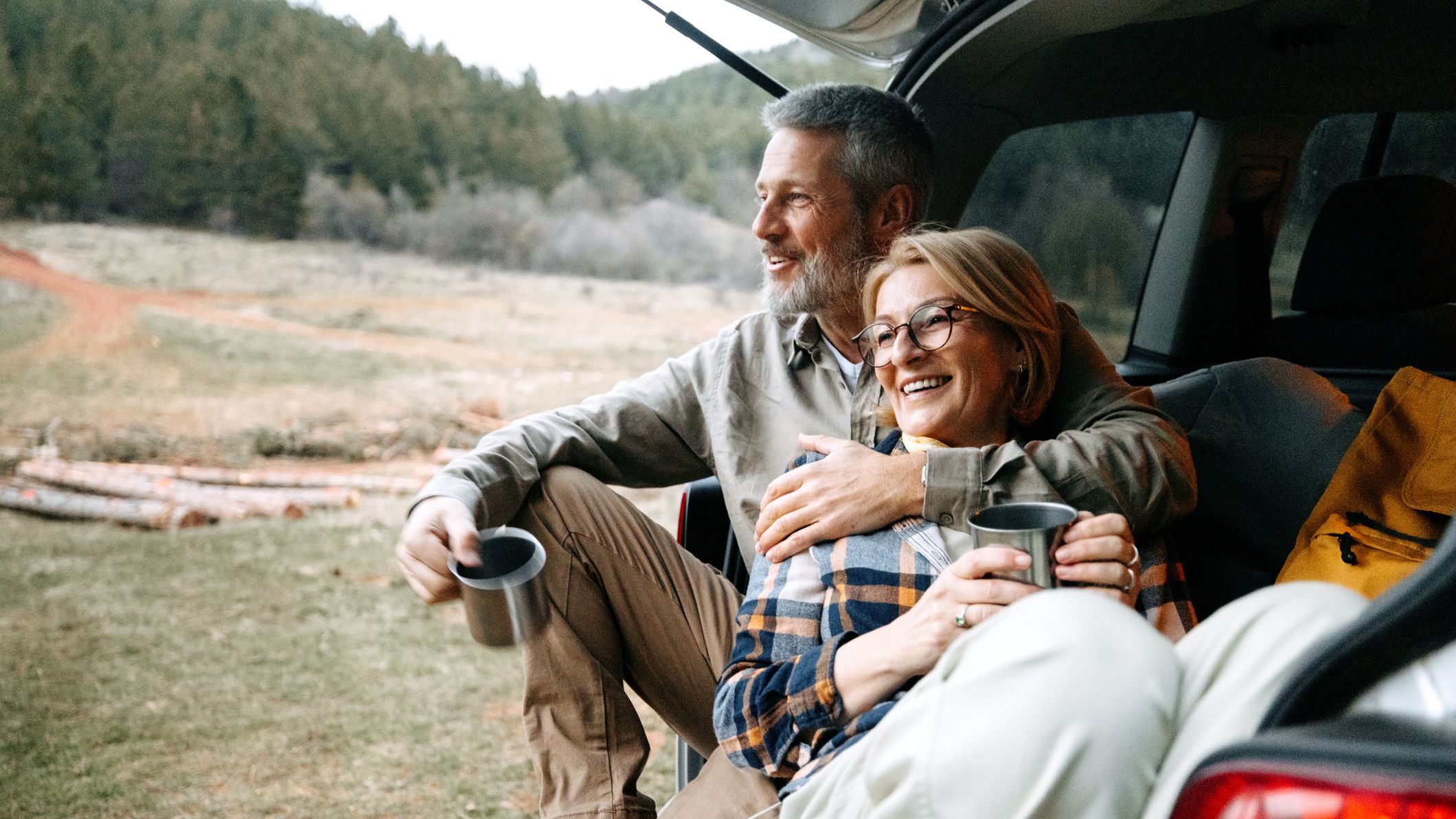 A mature couple takes a break after hiking by enjoying a hot drink by a car trunk in nature.