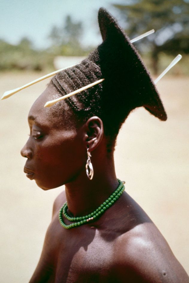 A dark-skinned woman looks to the left, her black braids wrapped around her conical shaped head, with a flat circular ring in the back and two wooden sticks poking through her hair. She stands in front of a tan background with blurred trees behind her