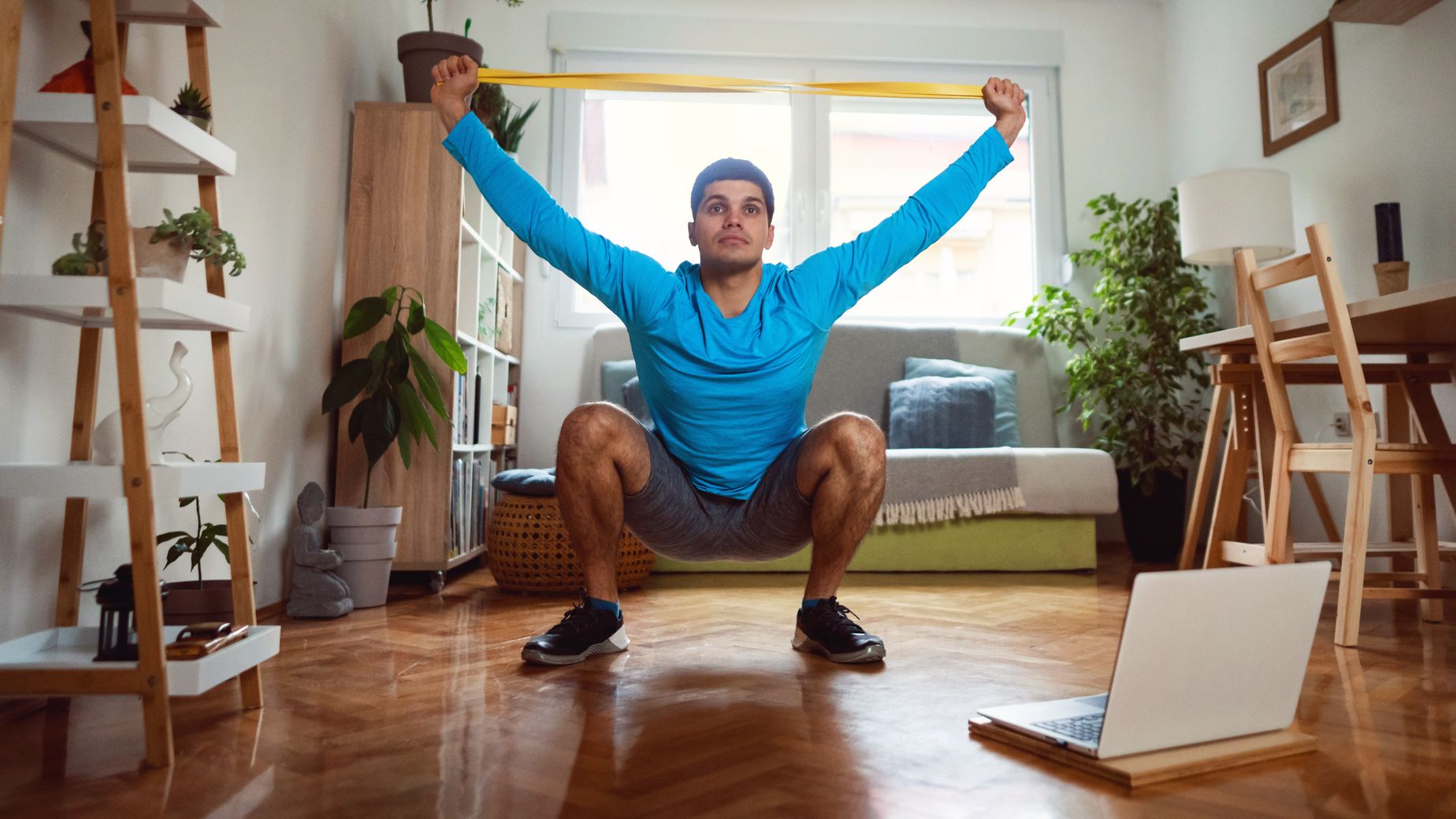 Man in sportswear in living room sits in a deep squat holding a resistance band in both hands above his head.