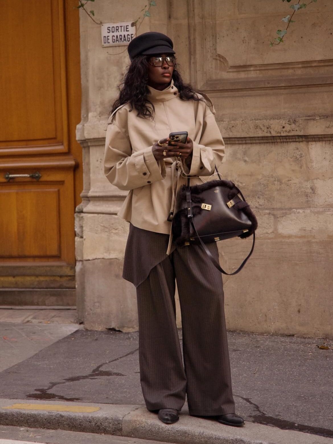 Fashion person Amy Lefevre wears a transitional winter to spring outfits 2026. She wears a tan funnel-neck cropped trench coat, brown trousers with a matching skirt and finishes her look with cowboy boots, the Ferragamo hug bag, a black bakerboy hat and large sunglasses. She is texting looking i the distance in a recent image taken from her Instagram.