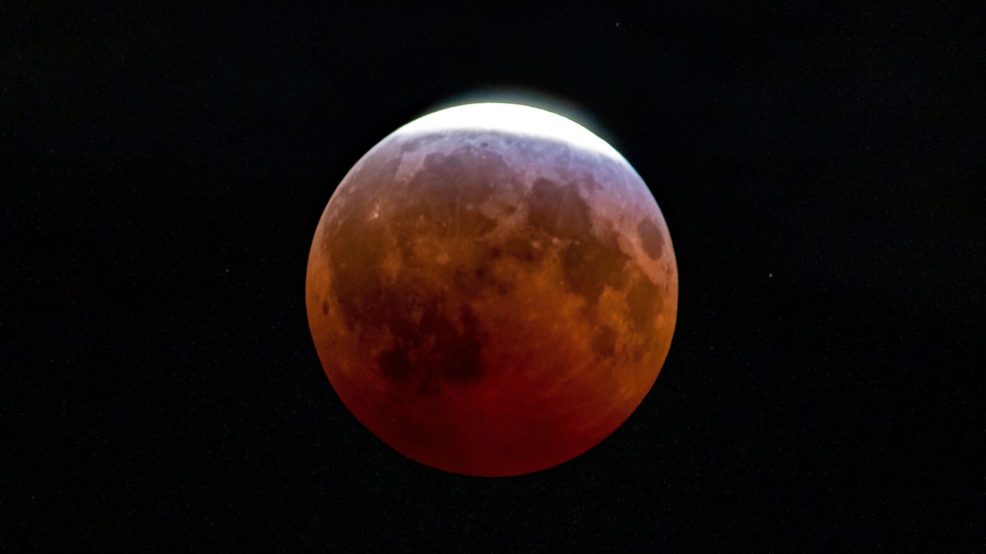 dark red moon with a bright patch of bright light as the remaining sliver of the lunar surface remains untouched by Earth's dark umbral shadow.