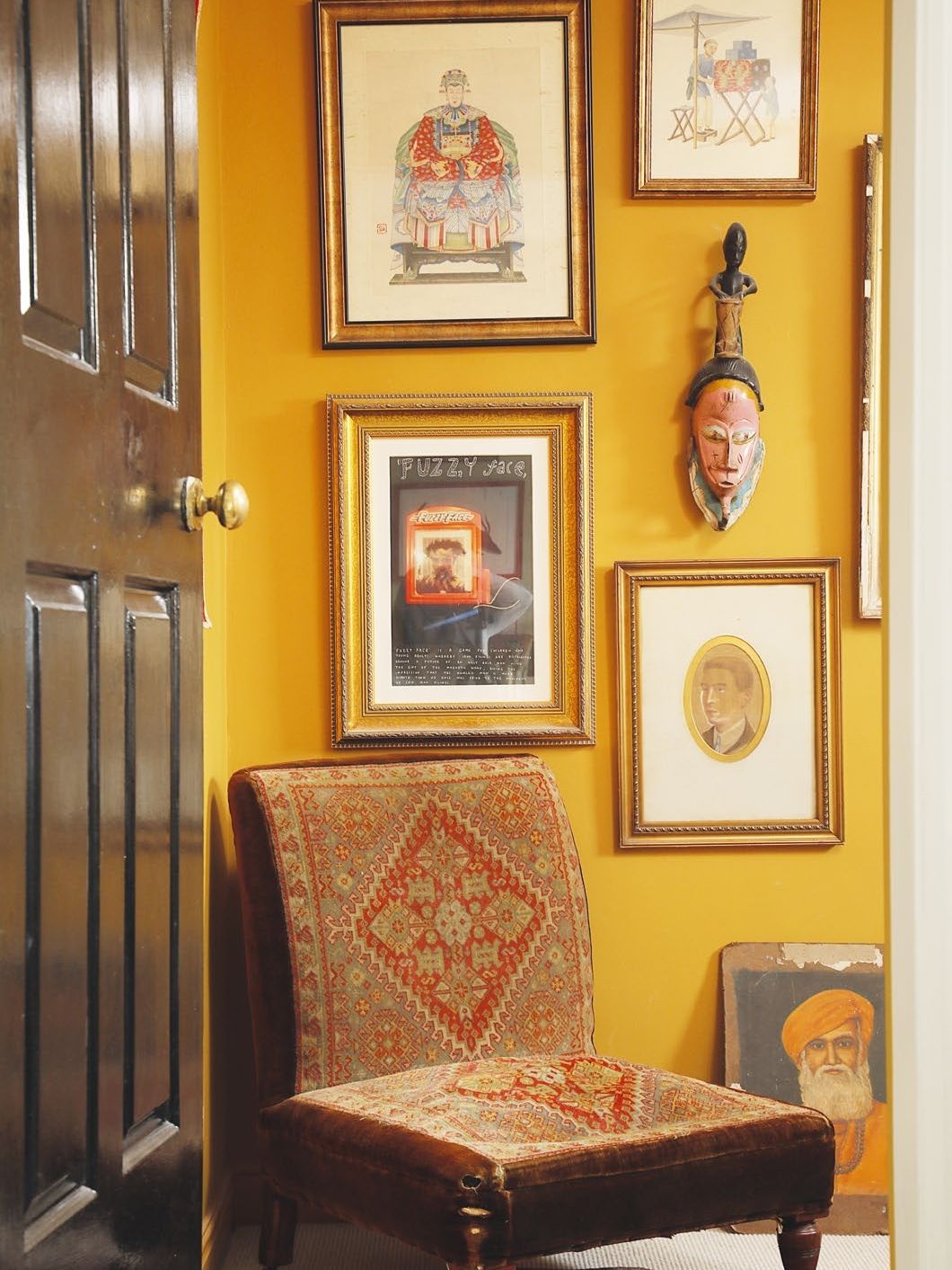 View into room with deep ochre painted walls, a low antique chais and mixed artwork on the walls