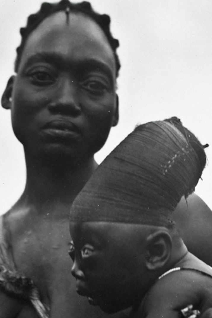 A black and white photo shows a dark-skinned woman with short hair holding her child, who is wearing a tight series of cloths wrapped around the top of its head to elongate its skull.