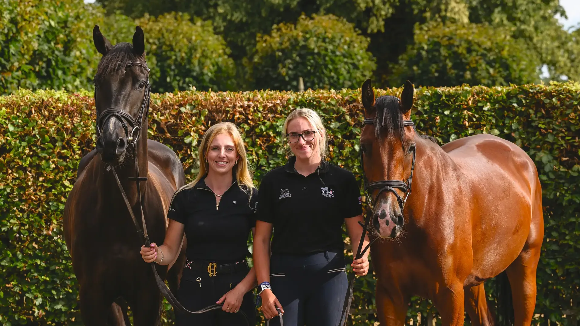 Cerys Macaulay and her groom holding two horses in front of a beech hedge