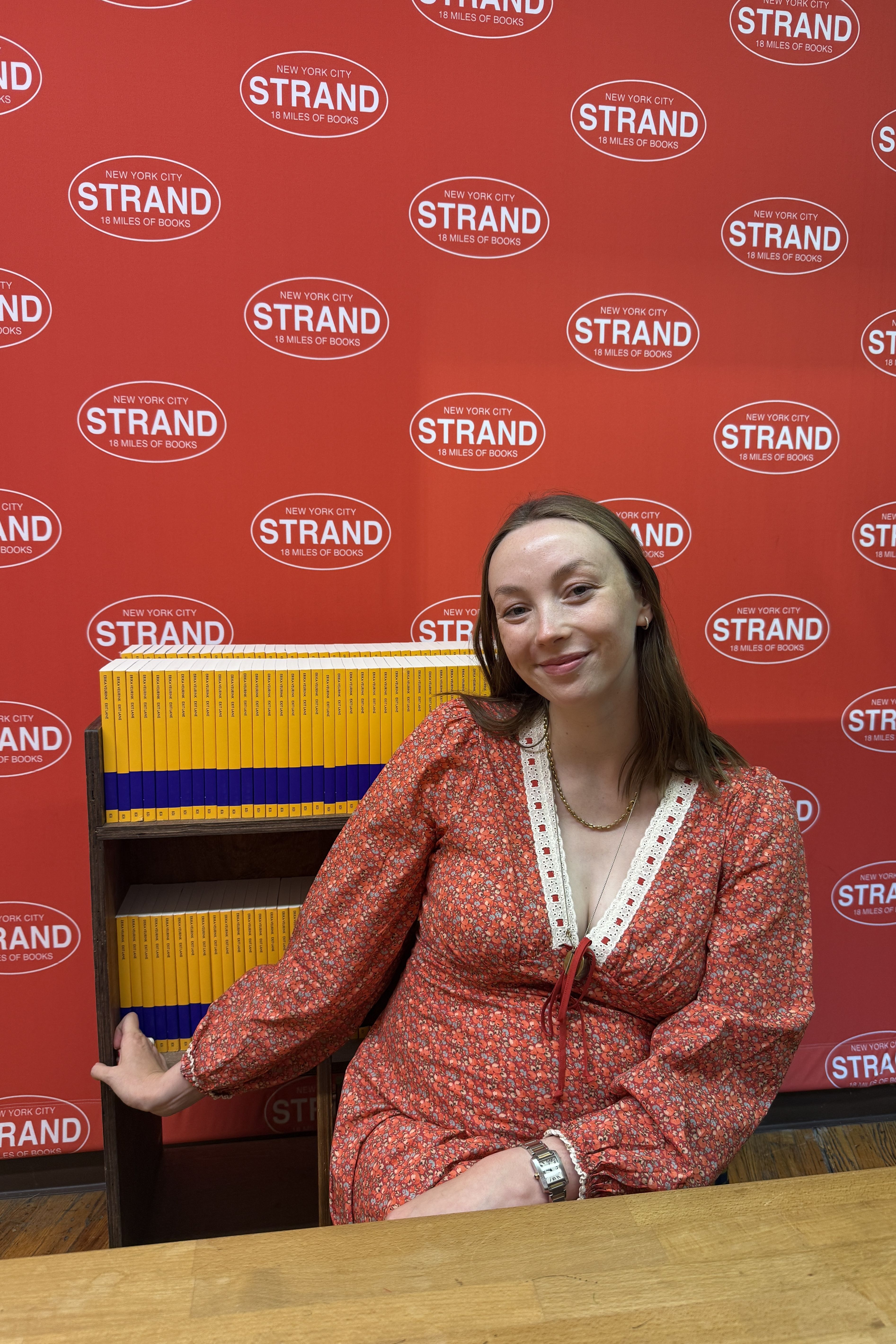 Erika Veurink sitting in front of a rack of her books at the strand