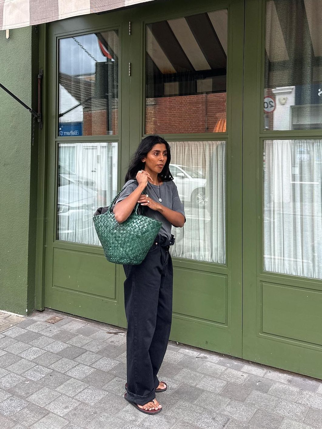 Woman wears gray t-shirt, black jeans, sandals and holds green dragon diffusion agola woven bag