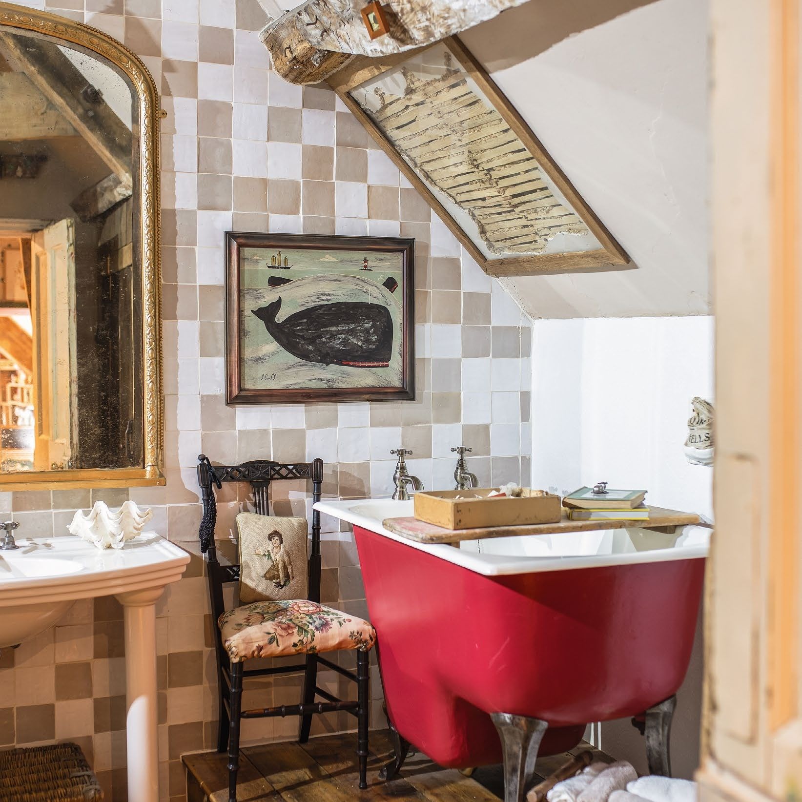 Cottage bathroom with red painted freestanding bath on wooden platform underneath eave with wooden beam. Wall covered in square neutral tiles.