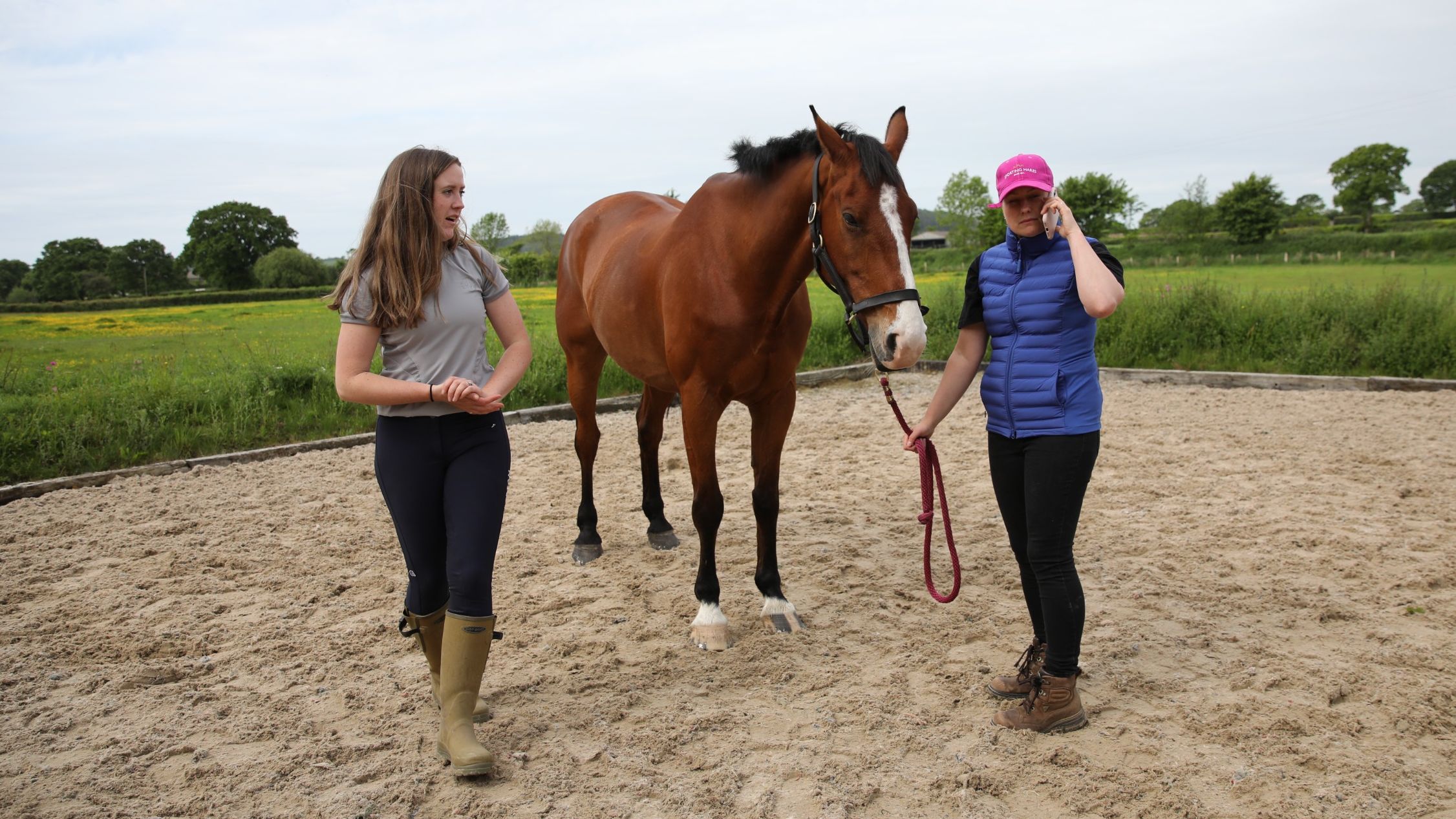 A bay horse stood in the corner of an arena with his handler and another woman; his handler is on the phone