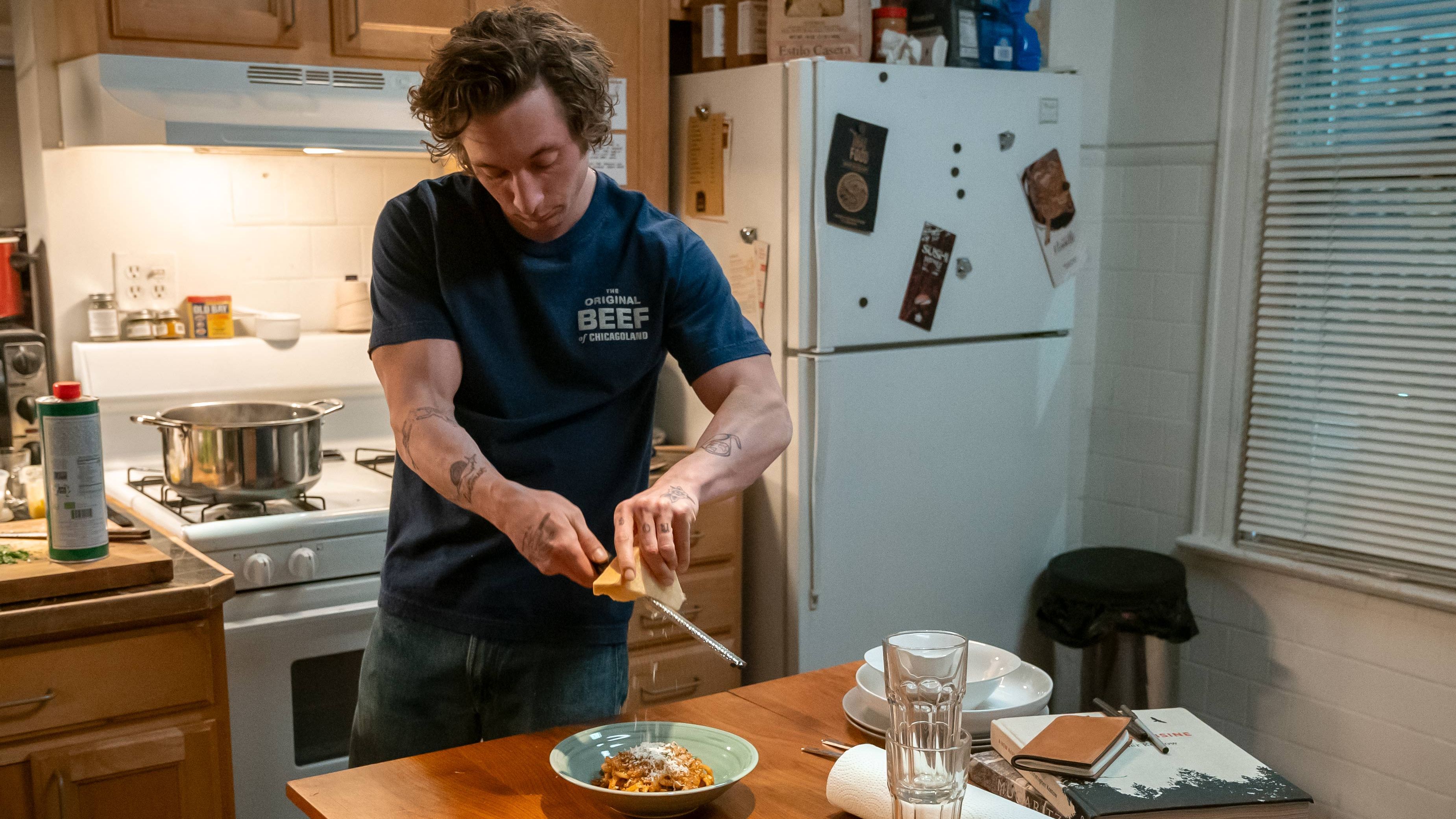 an image of Carmy (Jeremy Allen White) in The Bear grating cheese on top of a dish of pasta on a kitchen table at home