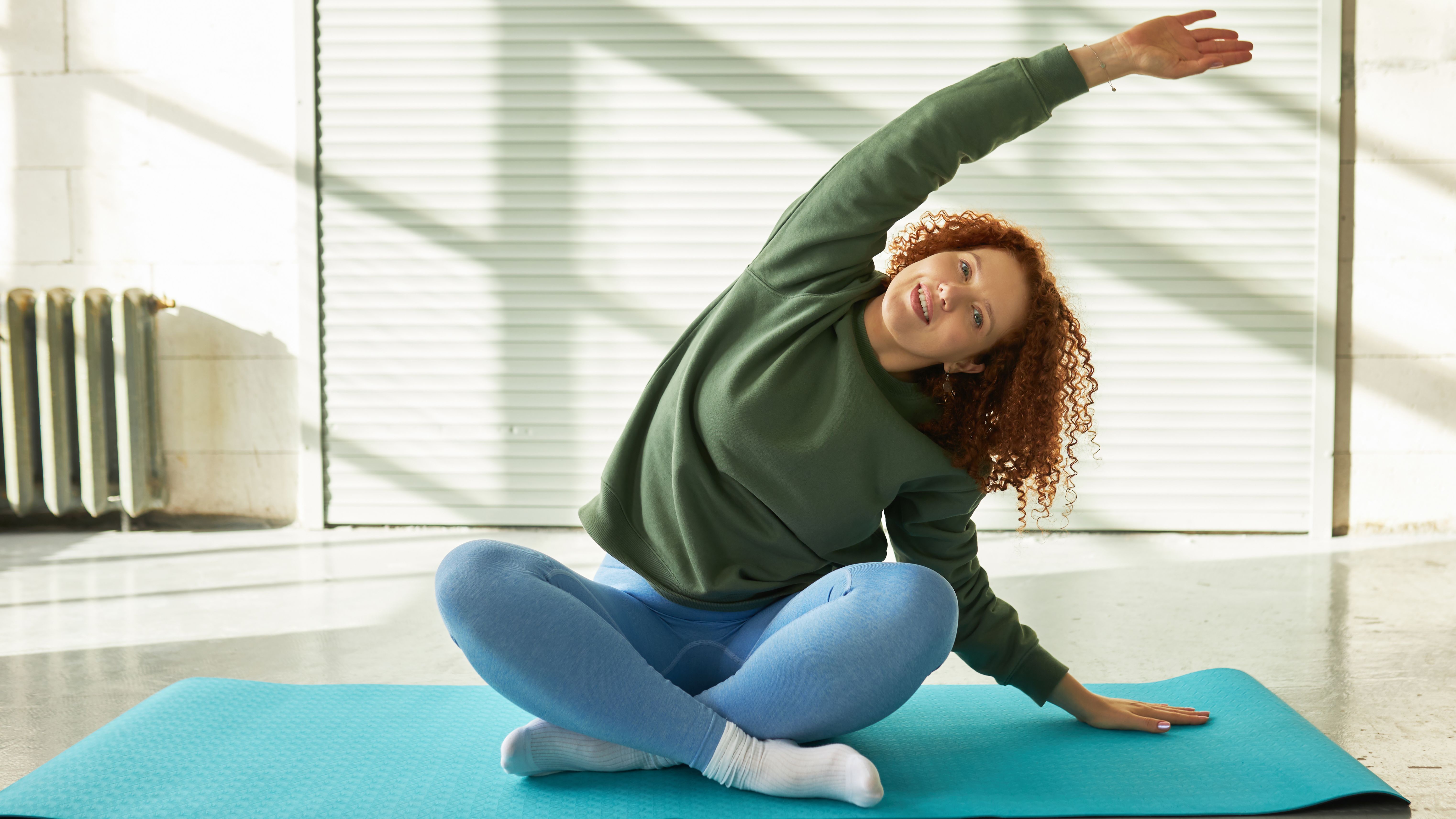 A woman sits cross-legged on a yoga mat, with her left forearm on the ground next to her, and her right arm stretched out up and her head to the left side. We see a white wall and radiator behind.
