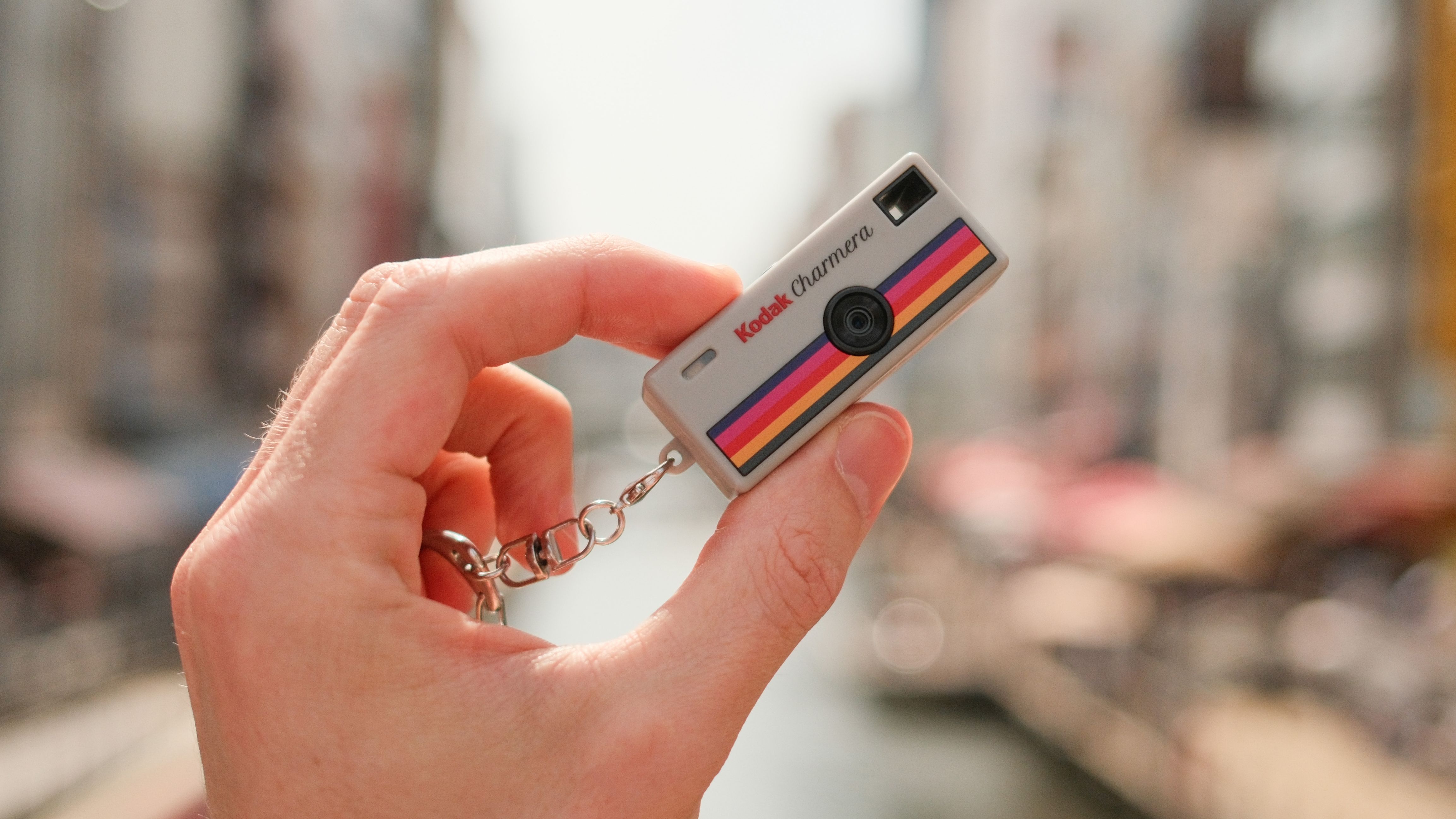 Hand holding a tiny Kodak Charmera keychain camera against a blurred canal background.