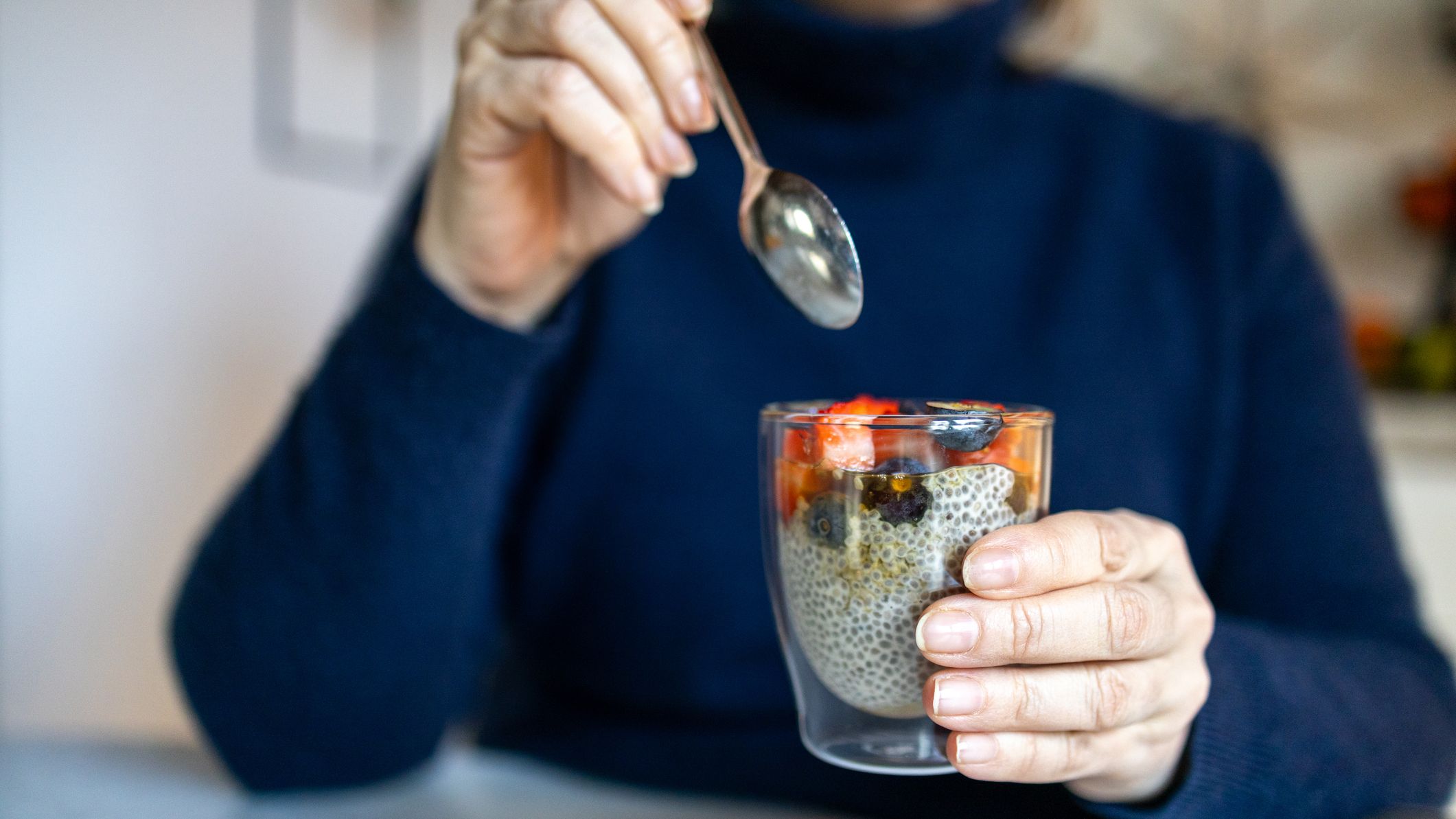 Woman holding a glass full of chia pudding and topped with fruit, a spoon is held above the glass