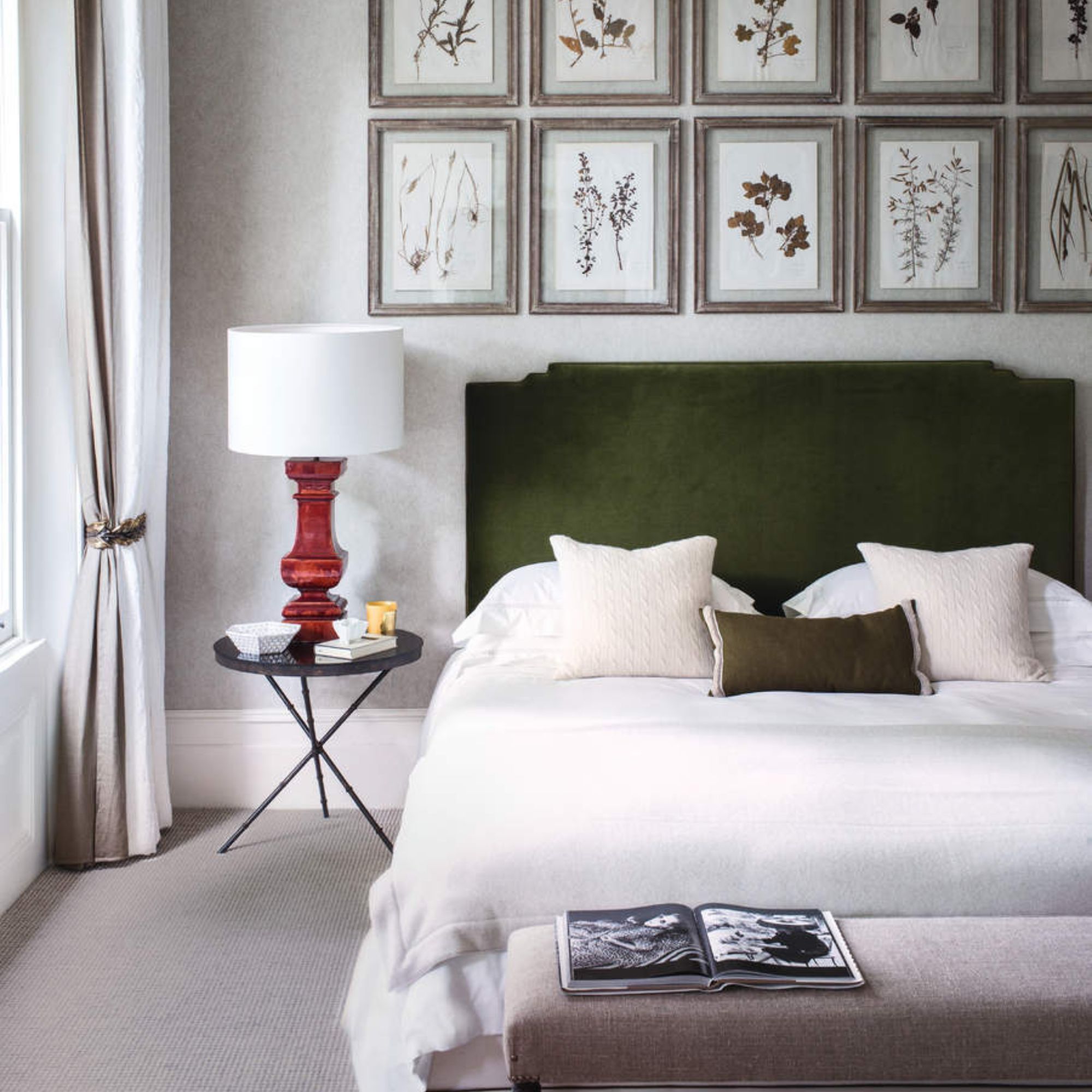 White and green bedding on a green bed frame against cream walls, covered in wall art; to the left a nightstand topped with a lamp; at the foot of the bed, a pink chaise lounge. 