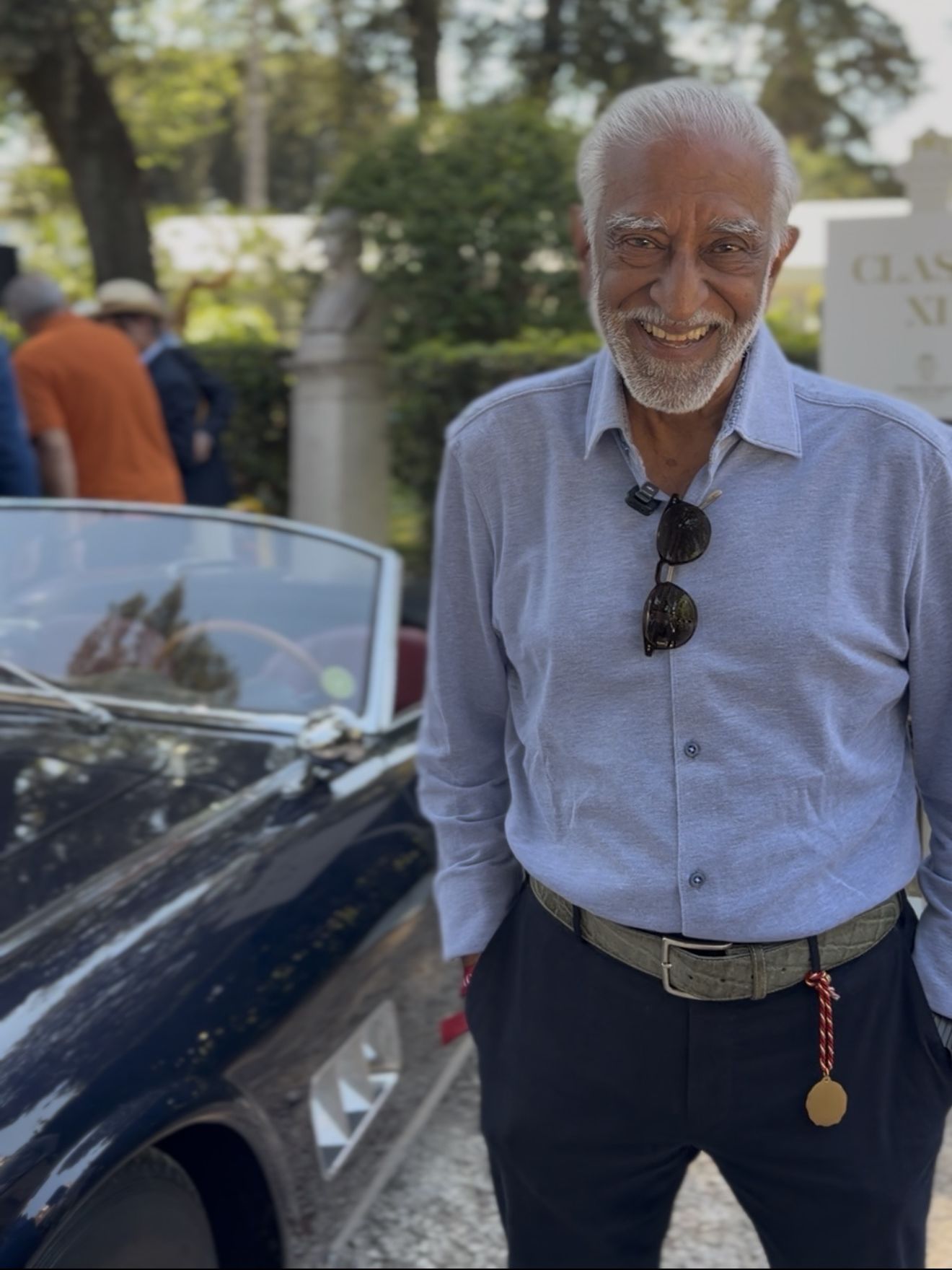 A smiling man stands beside a dark blue vintage convertible at Anantara Concorso Roma, with classic cars and concours guests gathered among trees in the background.