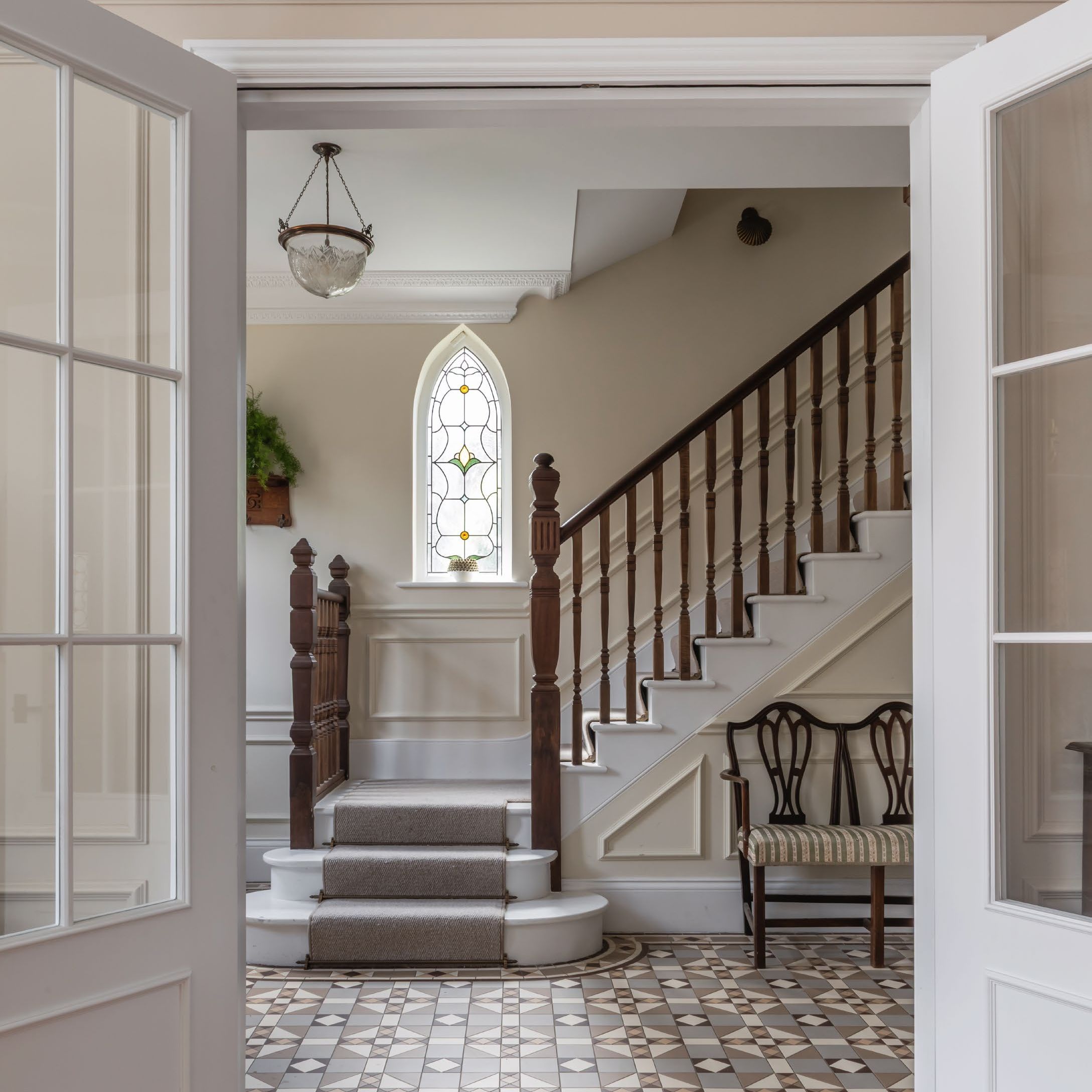 View through double doors into traditional hallway with encaustic floor tiles, wooden bannisters and small arched stained glass window