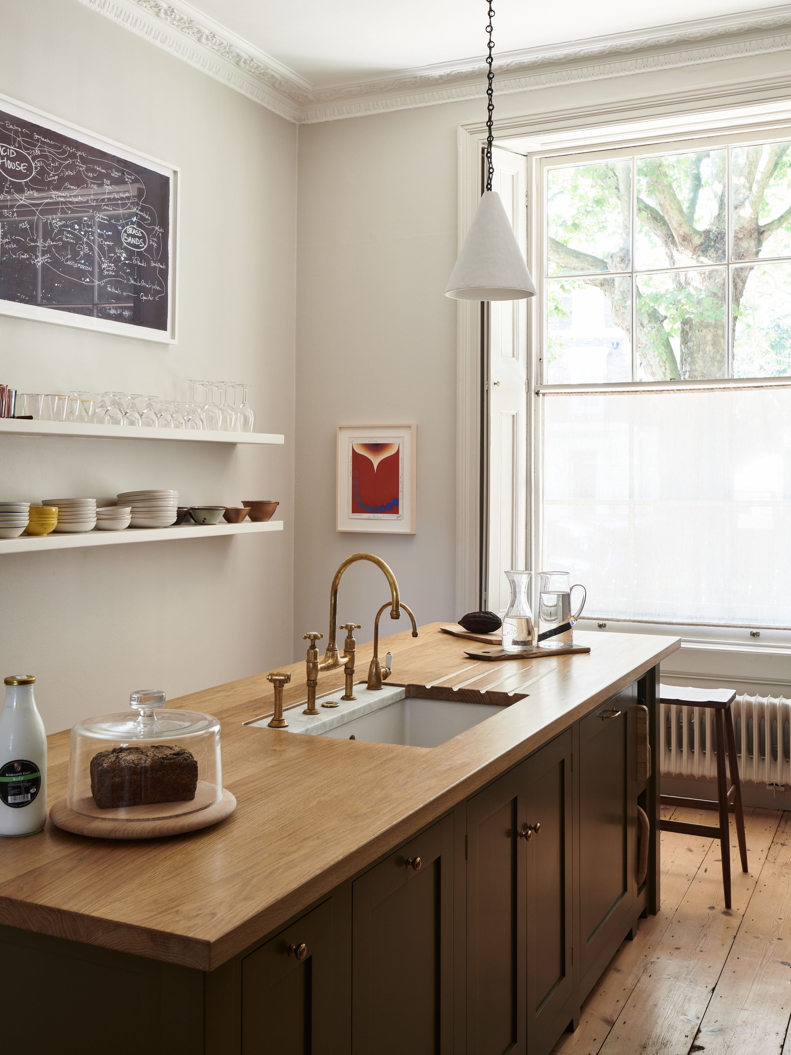 kitchen with wooden topped island with sink, open plan shelving on one wall, window, stool, wooden floor, pendant light, artwork